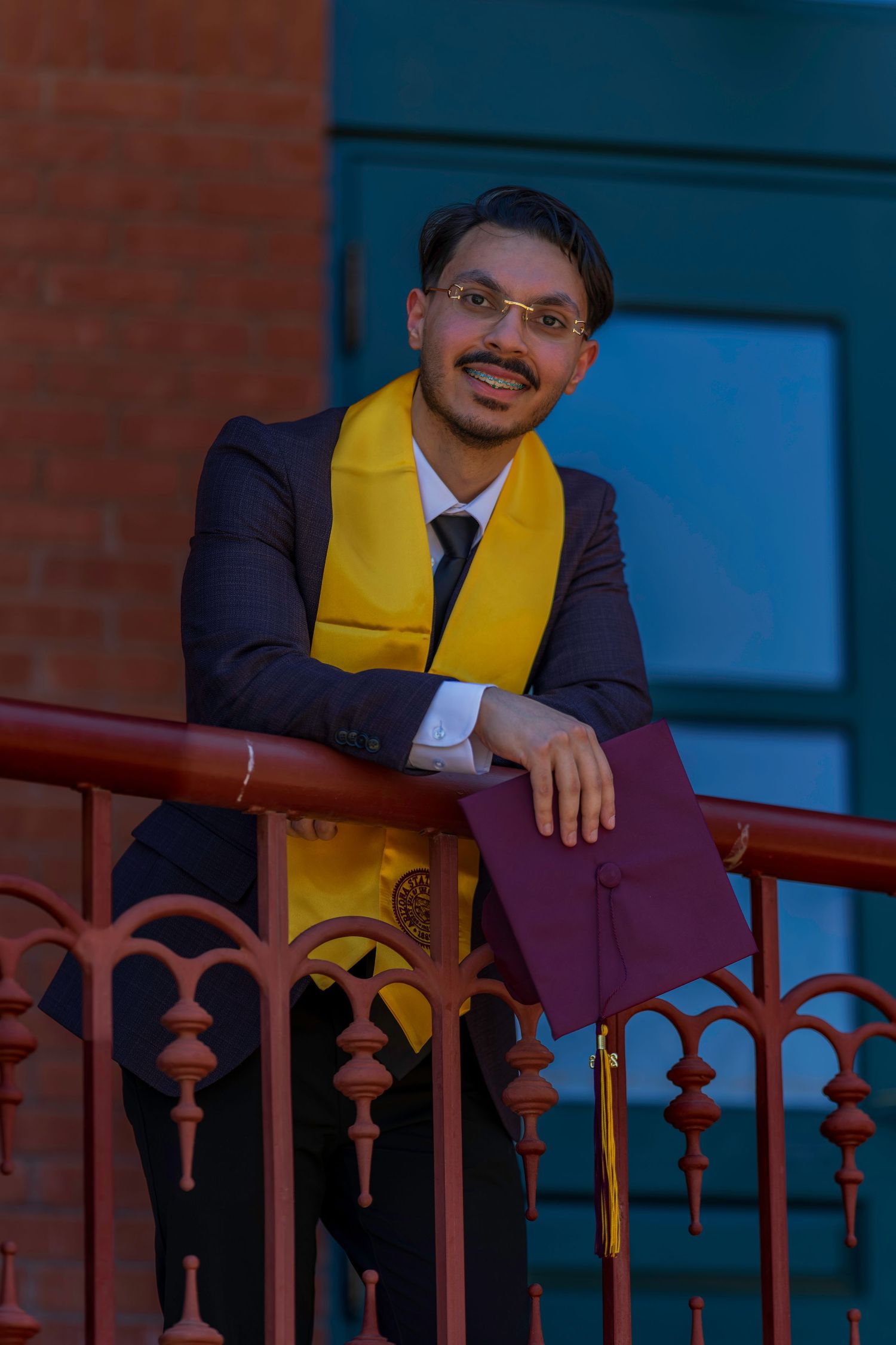 Smiling graduate in maroon suit and gold stole leans on ornate railing holding diploma.