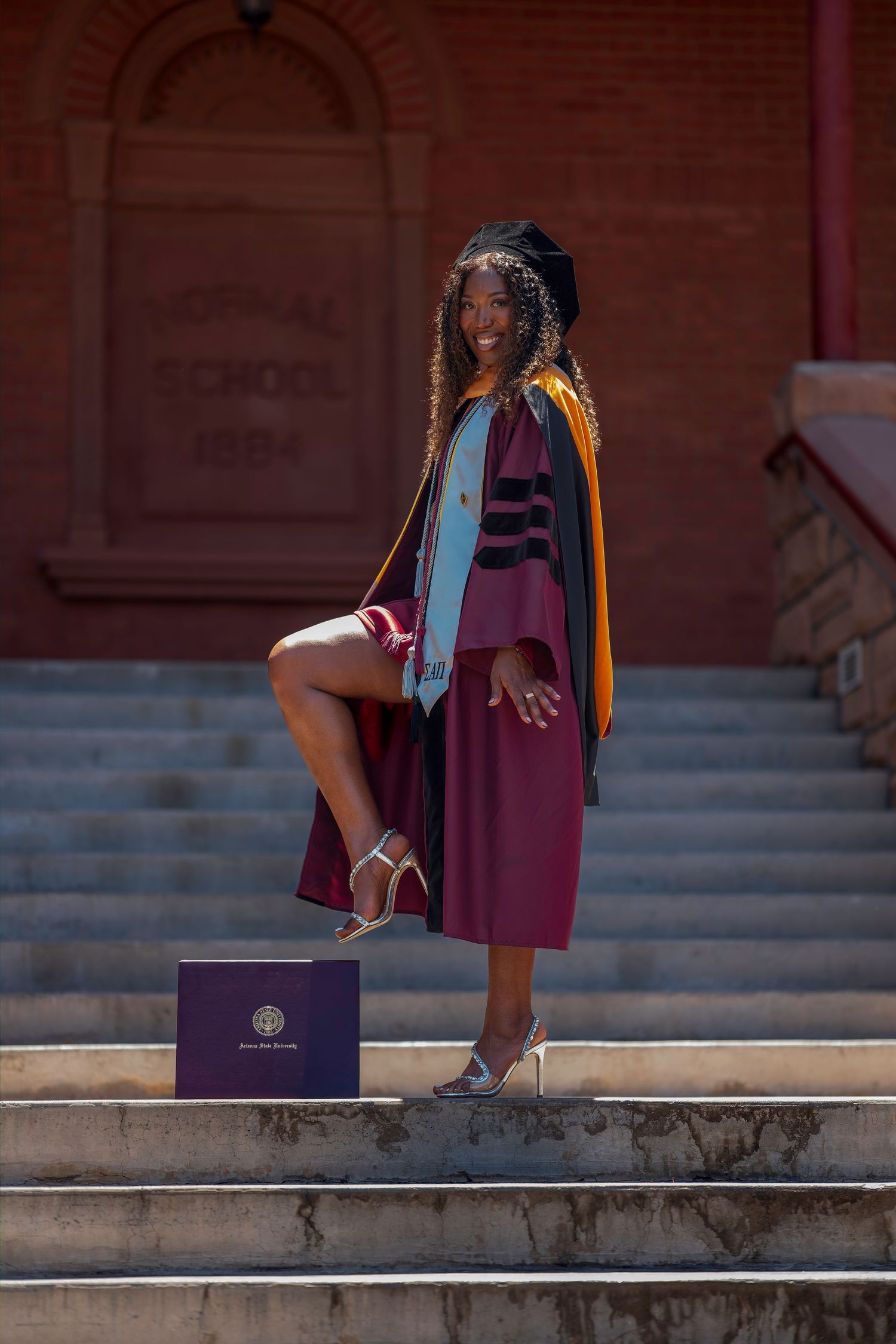 Smiling graduate in maroon cap and gown poses on steps with diploma on sunny day.