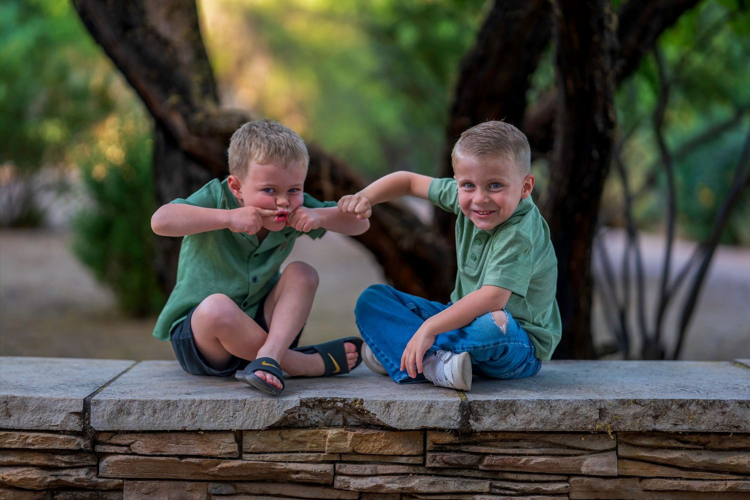 Two young boys in matching green shirts sit on a stone wall, laughing and pointing at each other outdoors.