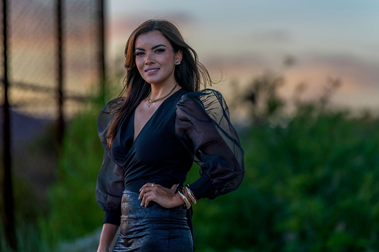 A smiling brunette woman in a black wrap top and skirt poses outdoors at sunset.
