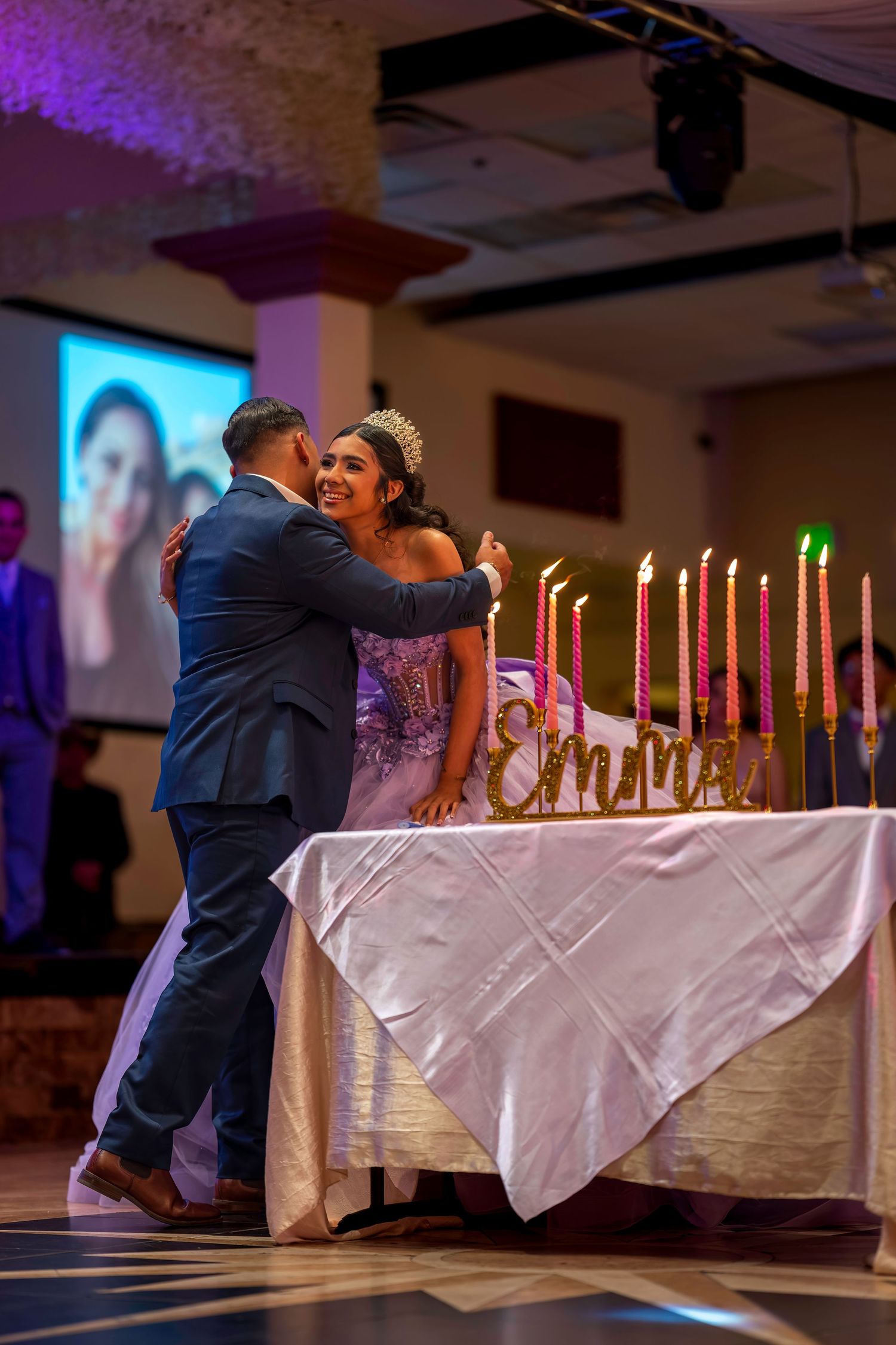 A couple embraces at a quinceañera celebration beside a candlelit table with a golden Emma name sign.