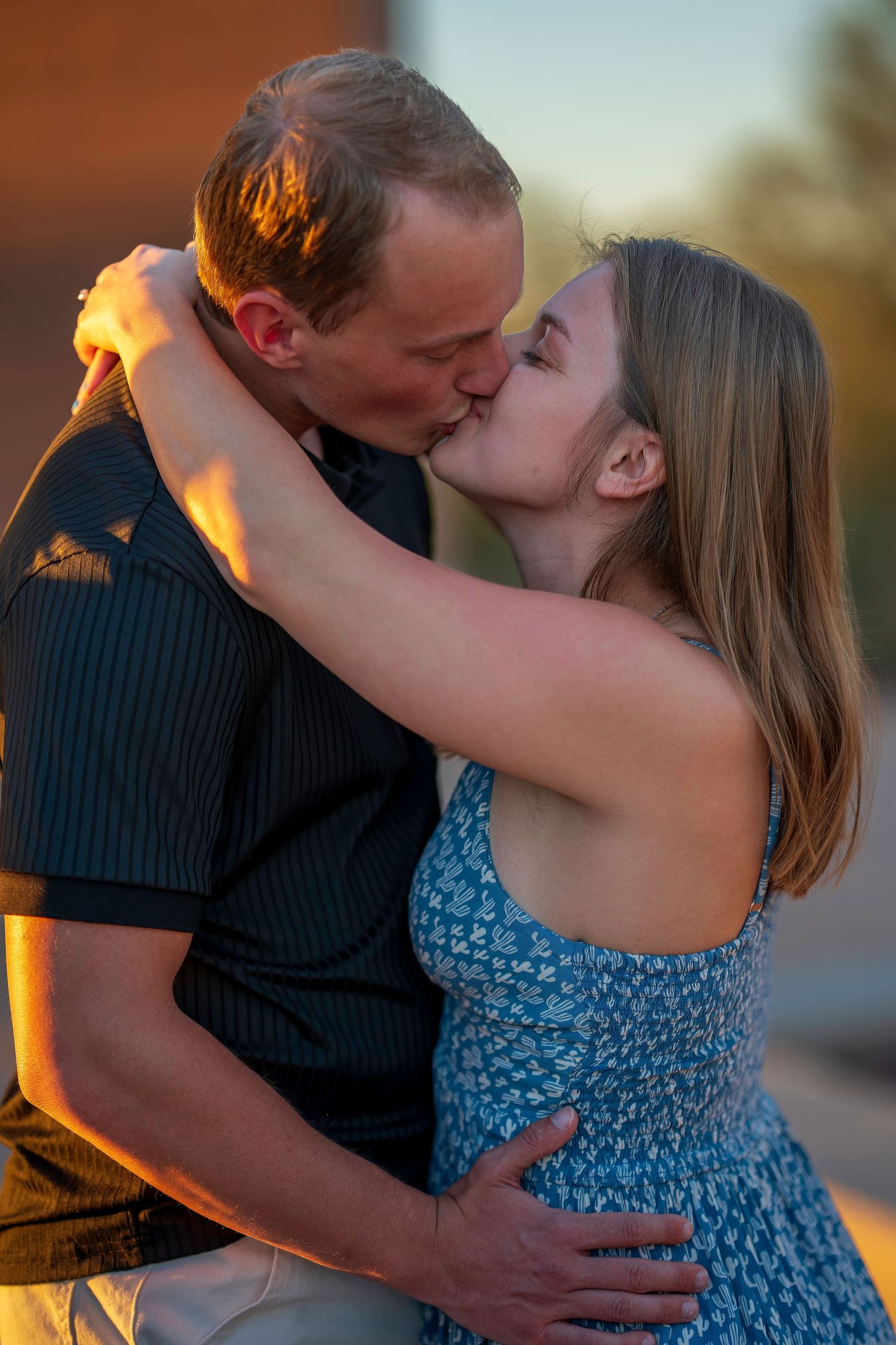 Couple sharing a passionate kiss outdoors during warm golden sunset light, woman in blue dress, man in black shirt.