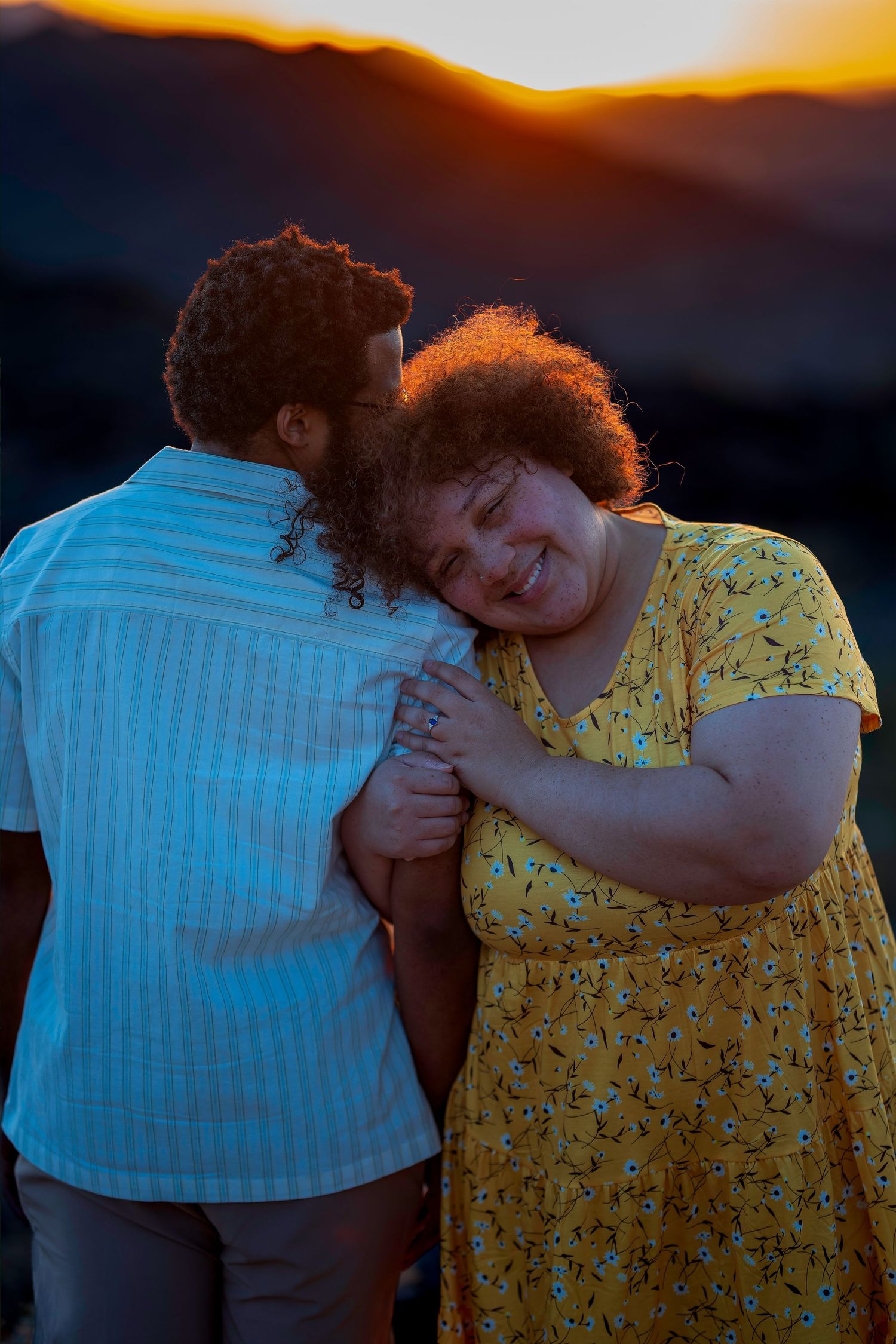 Couple embracing at sunset, woman in yellow floral dress smiling as she leans on mans shoulder.