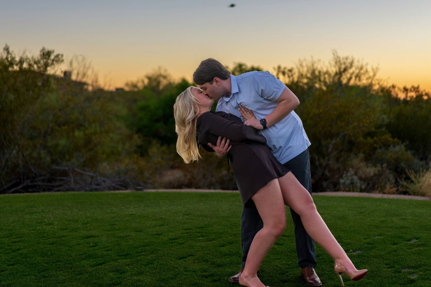 Romantic couple sharing a dip kiss on green lawn at golden sunset with desert landscape behind.