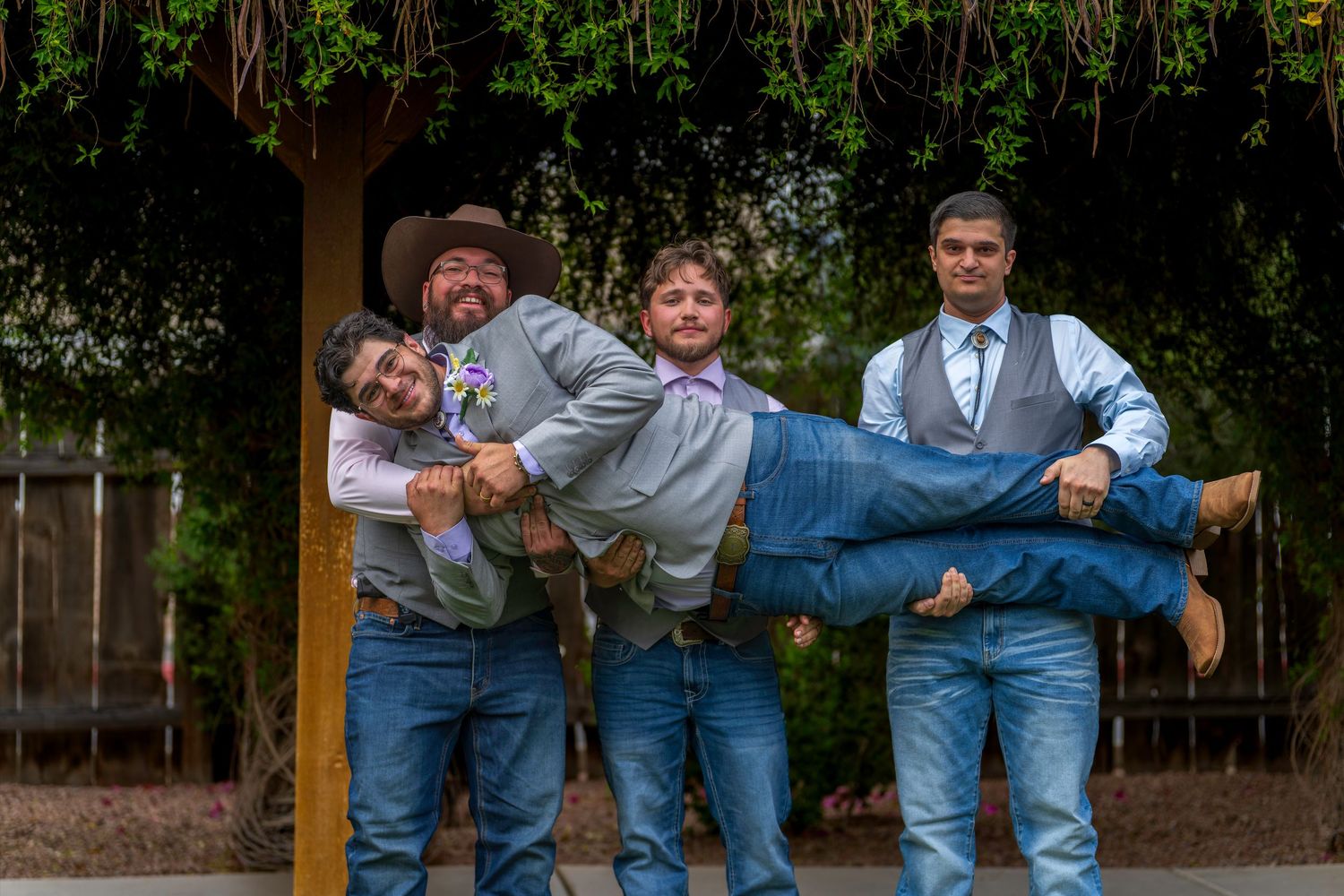 Four men in wedding attire joking around outdoors, with three holding one man horizontally in the air.