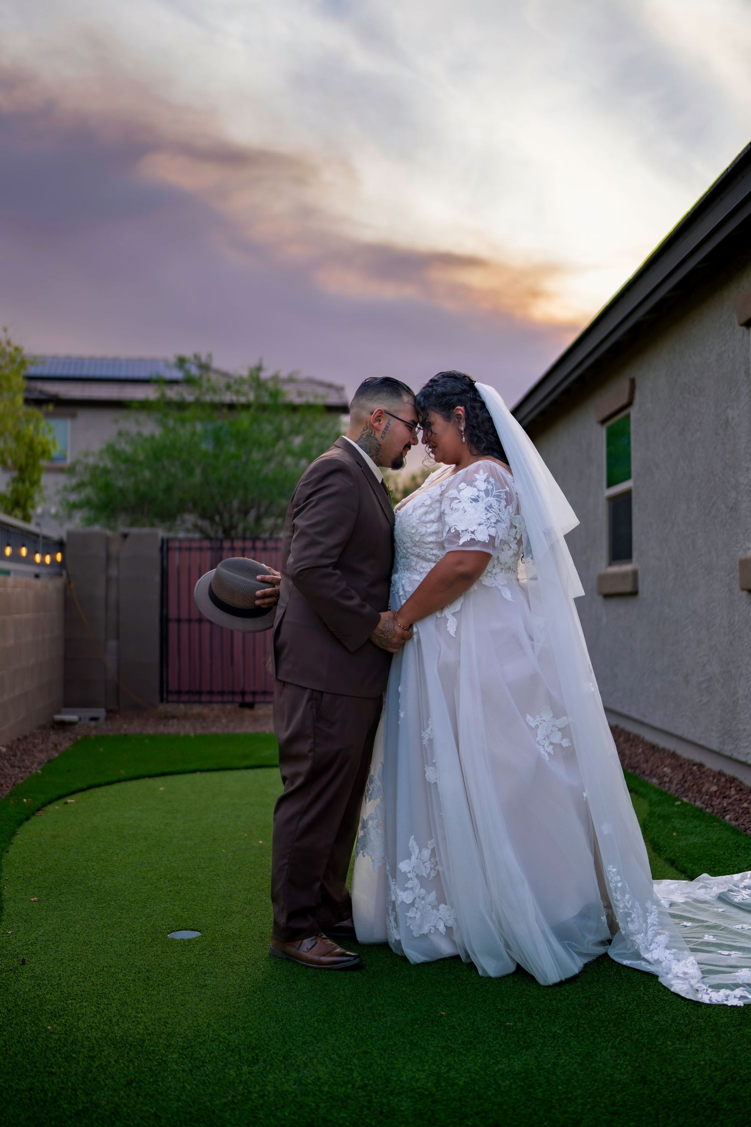 Bride and groom share a romantic moment on green turf at sunset, bride in flowing lace gown with veil.
