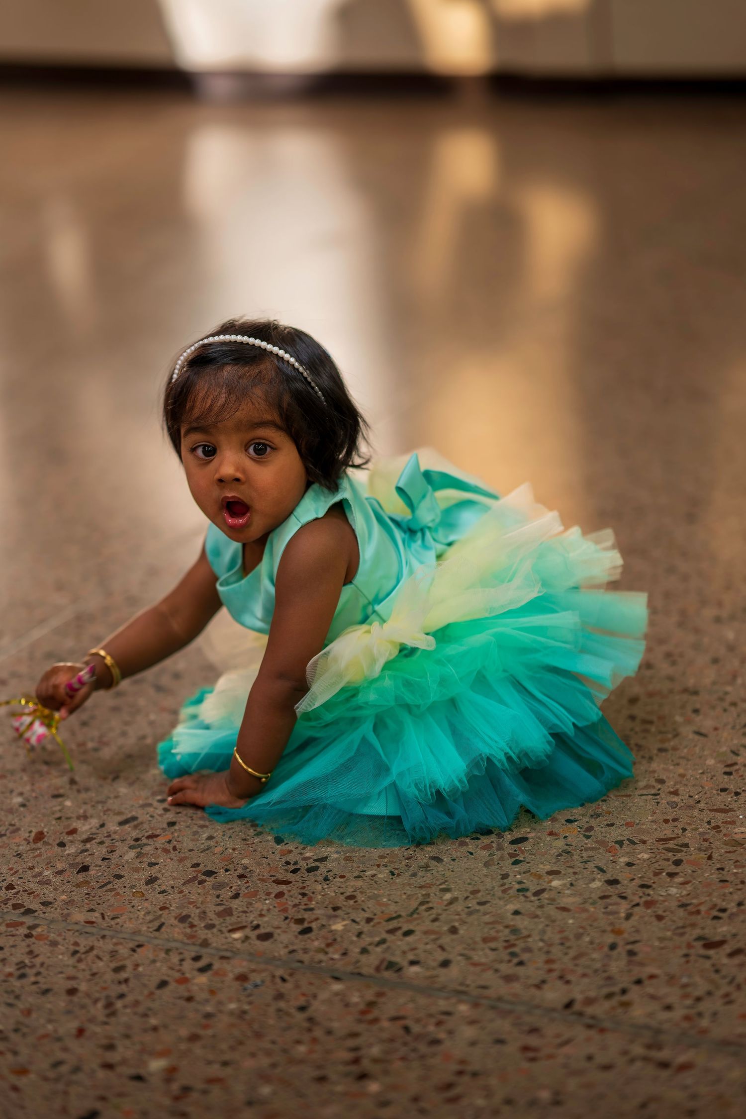 A baby girl in a teal tutu dress crawls on a marble floor wearing gold bangles and a headband.