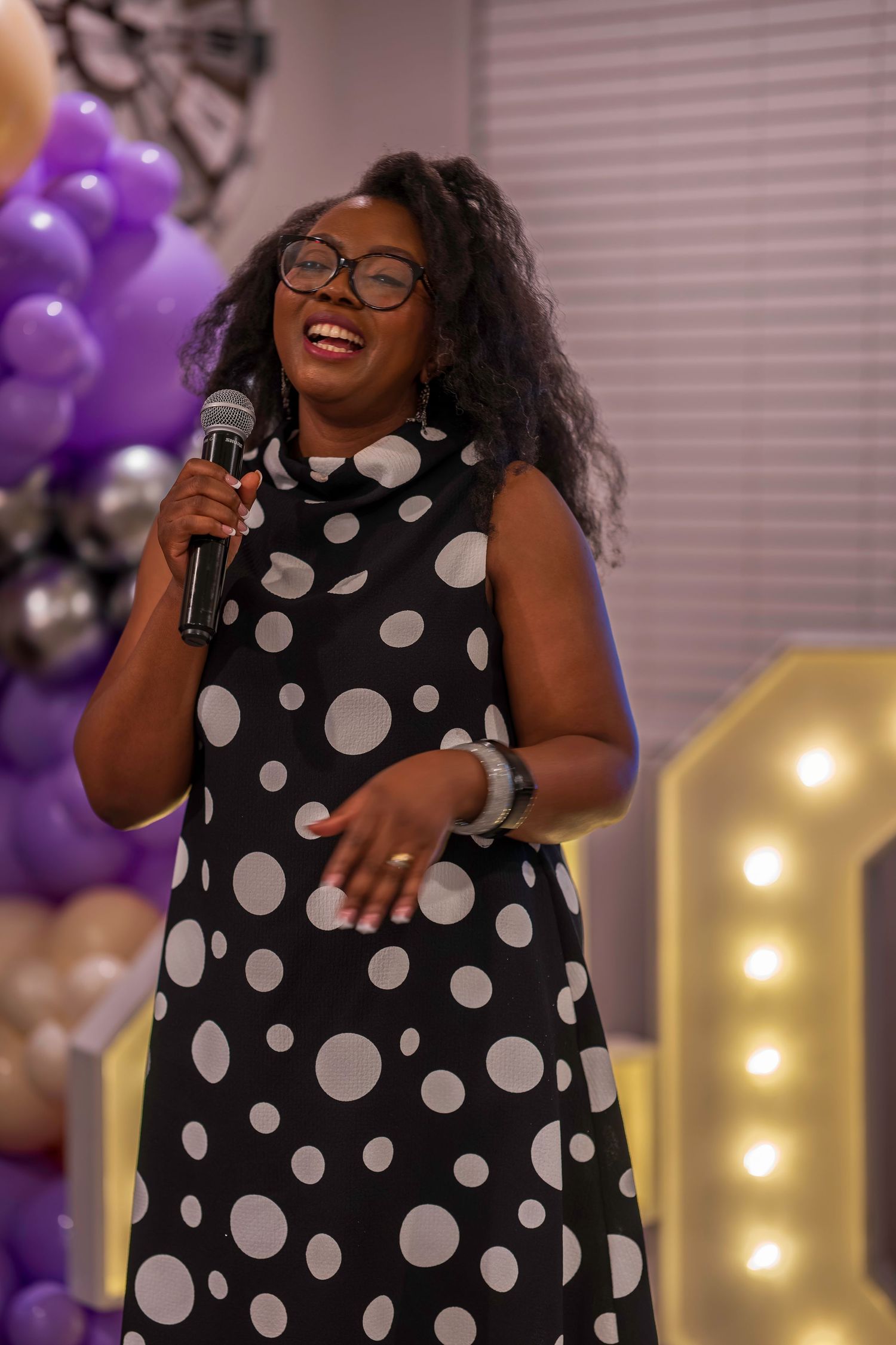 A smiling Black woman in a polka dot dress holds a microphone at an event with purple balloons.
