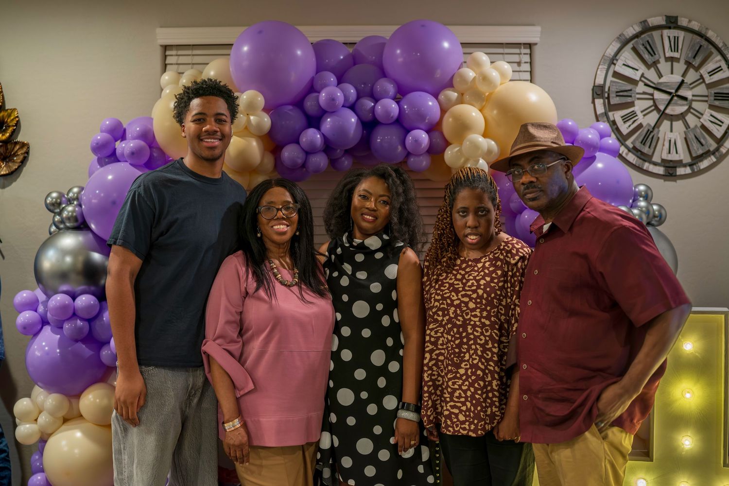 Group of five people posing together in front of purple and gold balloon arch at a festive celebration.