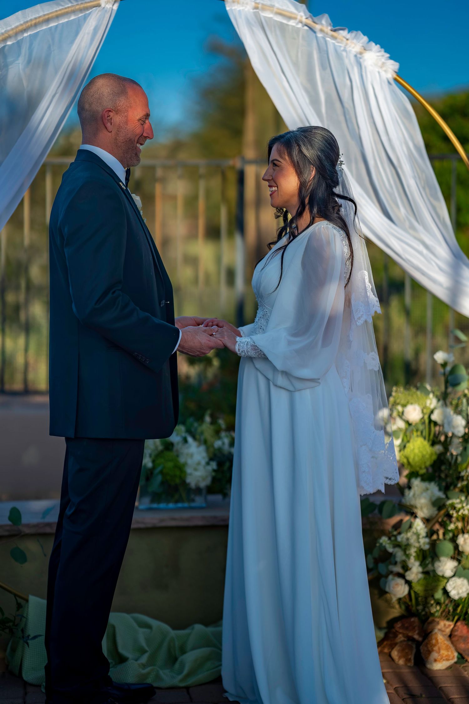 Bride and groom exchange vows under a draped arch adorned with white flowers at outdoor wedding ceremony.