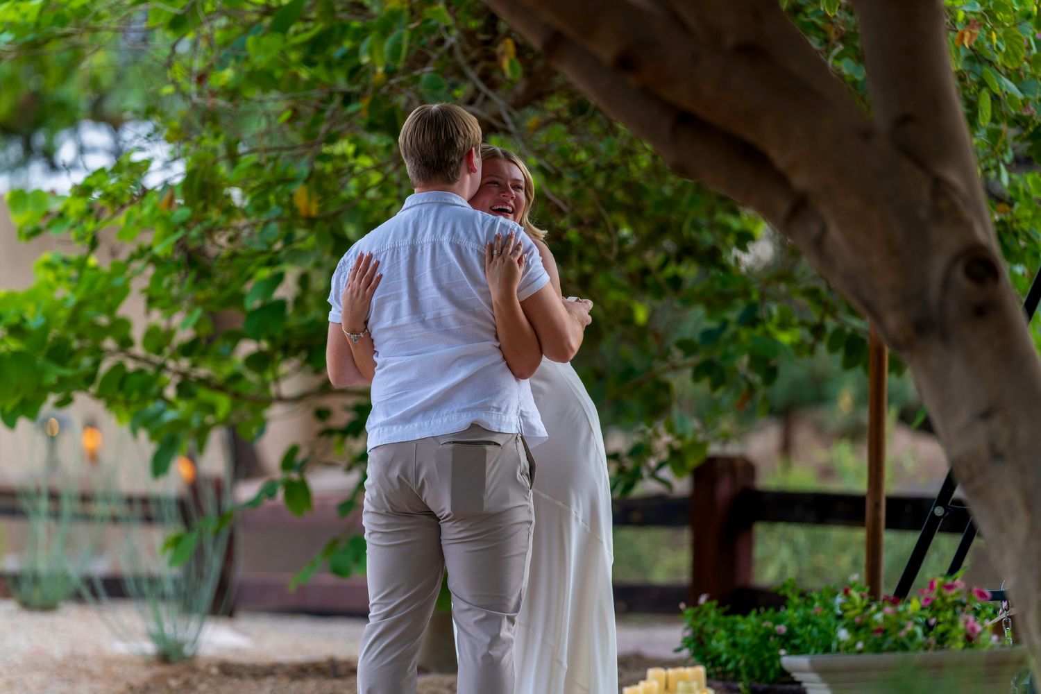 Couple embracing tenderly under a lush green tree in an outdoor garden setting during a romantic moment.
