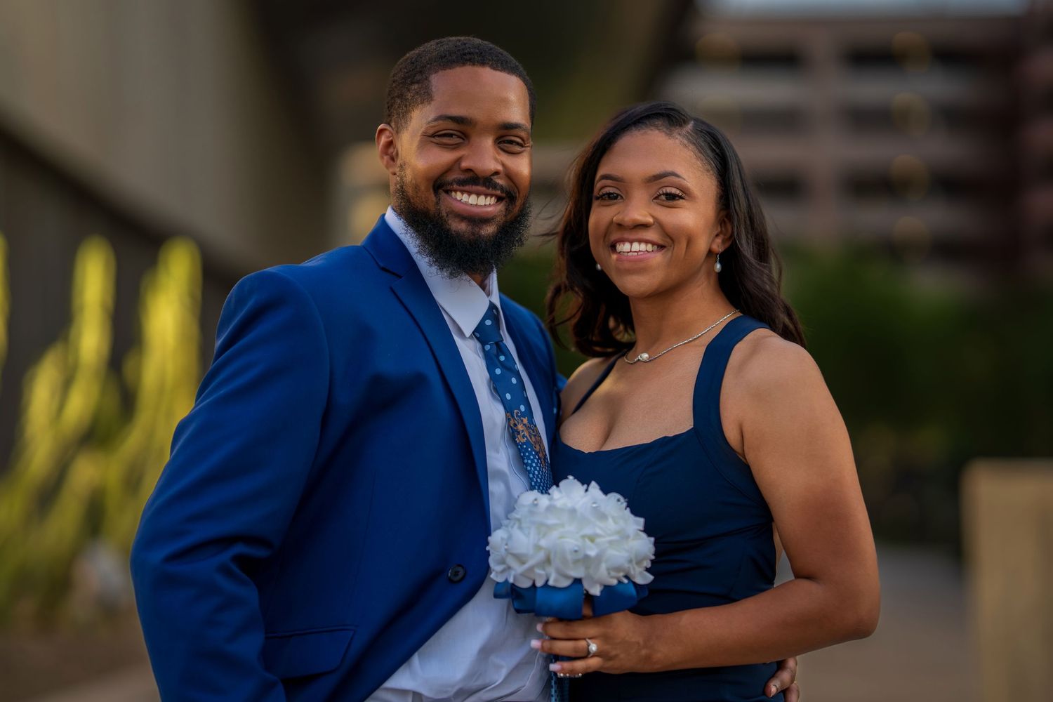 Smiling couple in matching navy blue outfits posing outdoors, woman holding white hydrangea bouquet.