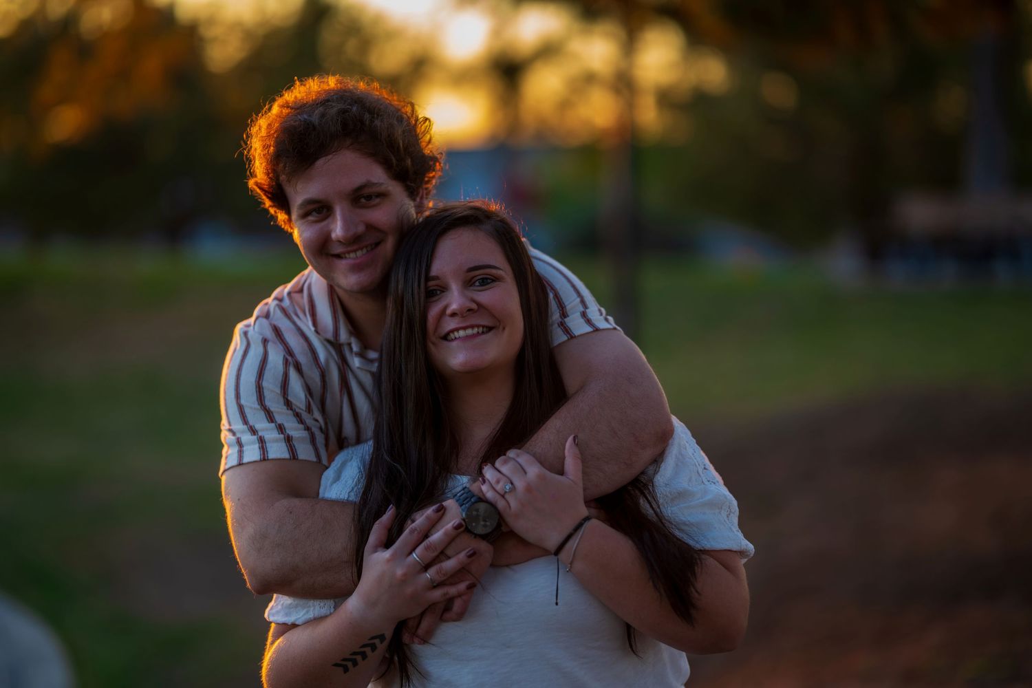 Happy couple embracing and smiling in a park during warm golden sunset light, man hugging woman from behind.