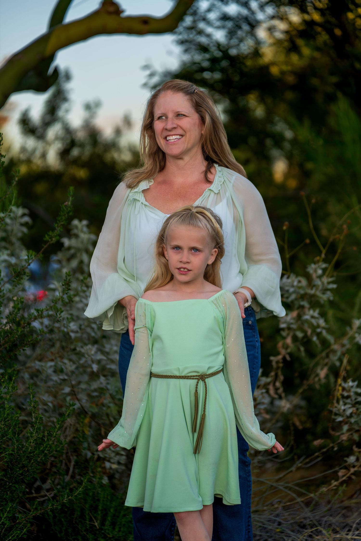 Woman and young girl in matching green and white outfits posing together outdoors among lush greenery.