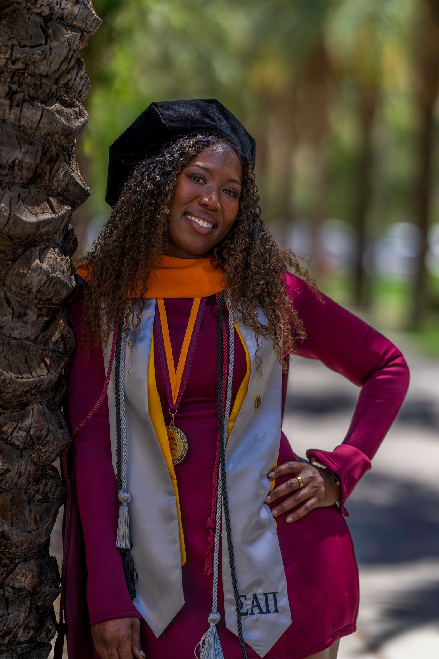 Smiling woman in maroon graduation gown, black cap, gold medal, and white stole poses by palm tree outdoors.