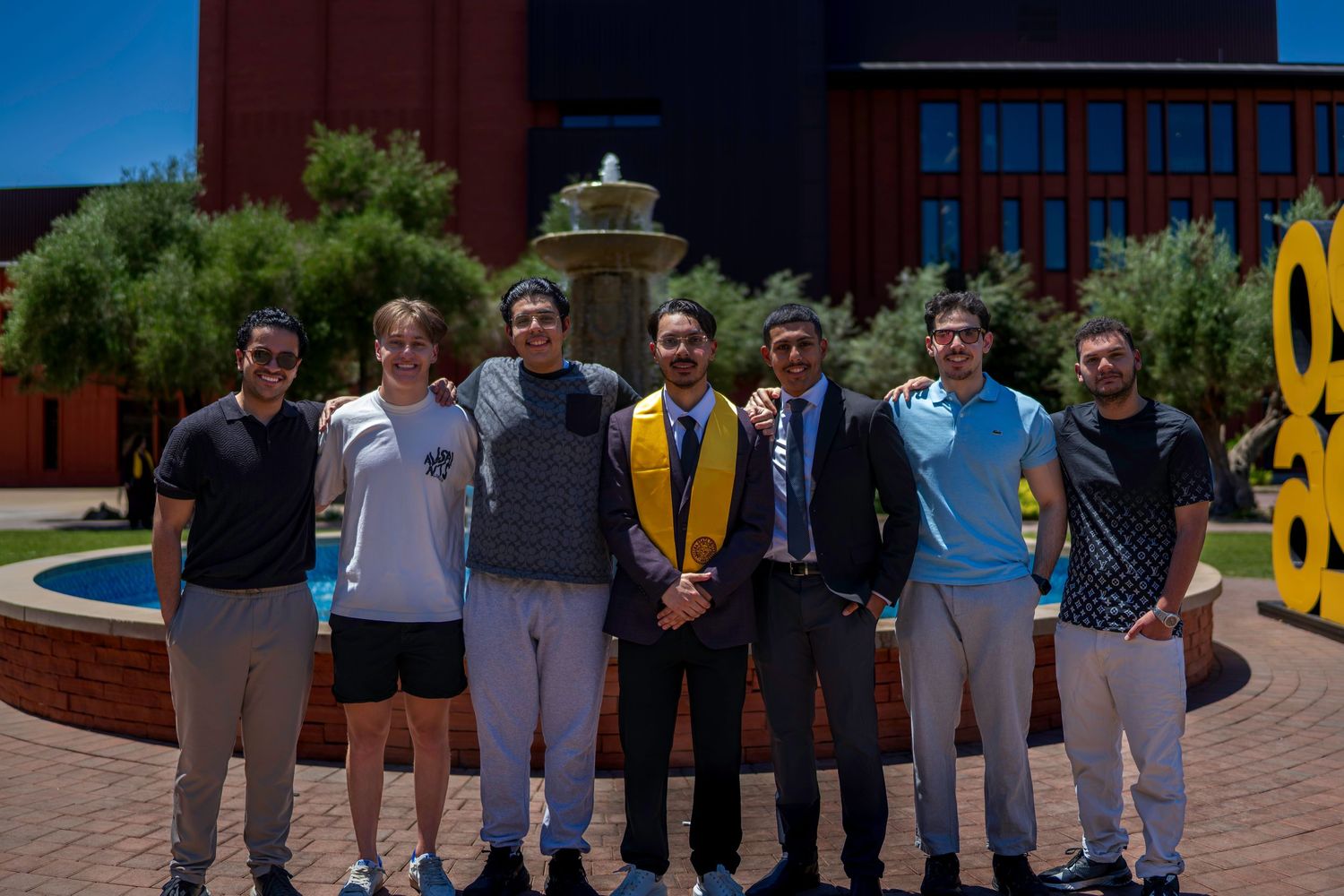 Group of seven young men posing outdoors near a fountain, one wearing graduation regalia with gold stole.