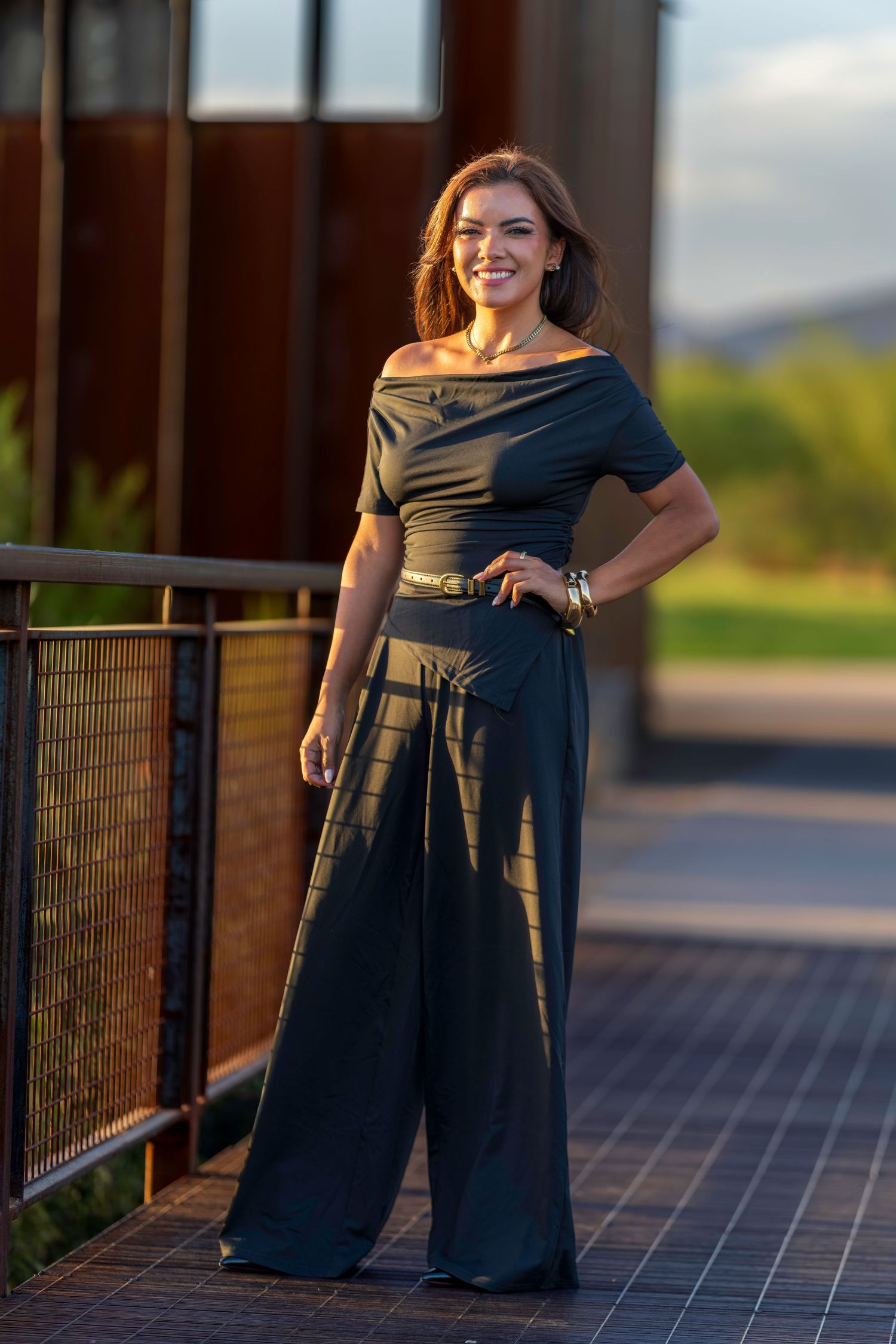Woman in elegant black off-shoulder jumpsuit with wide-leg pants smiling outdoors at golden hour.