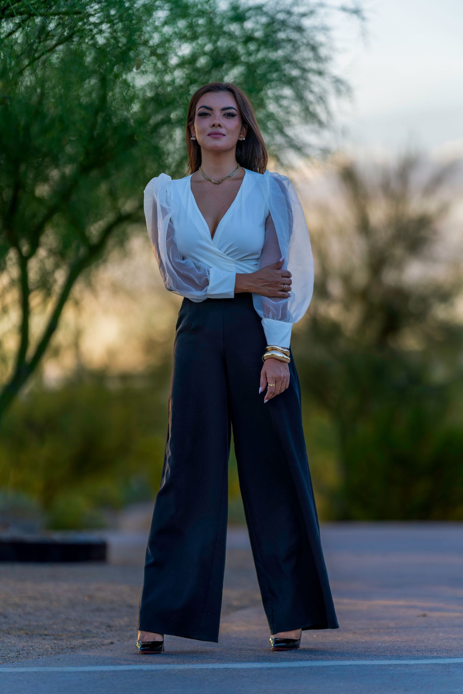 Woman in white wrap blouse and black wide-leg pants poses outdoors at sunset with green trees in background.