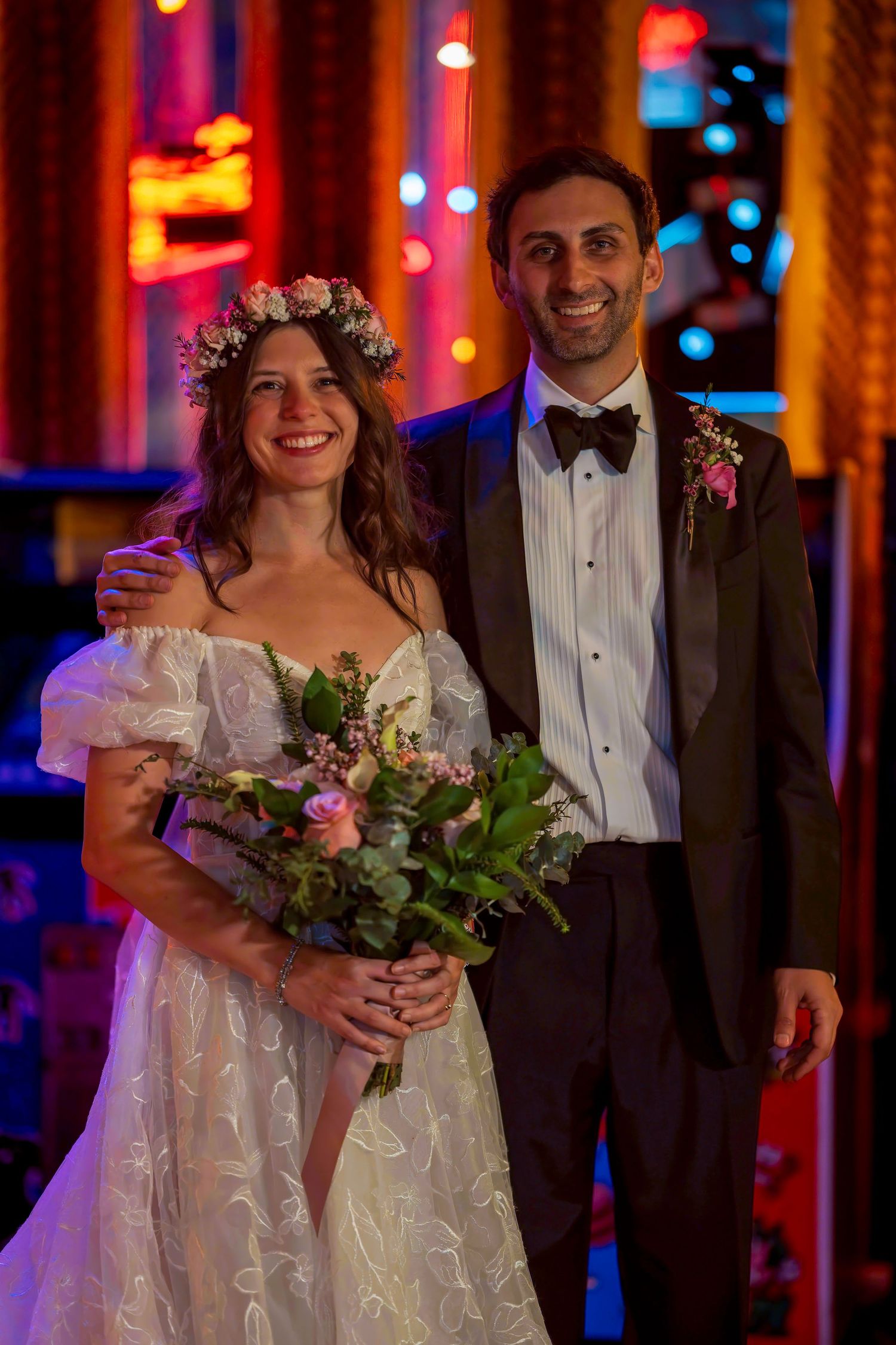 Smiling bride in white lace gown with floral crown holding bouquet beside groom in black tuxedo at wedding reception.