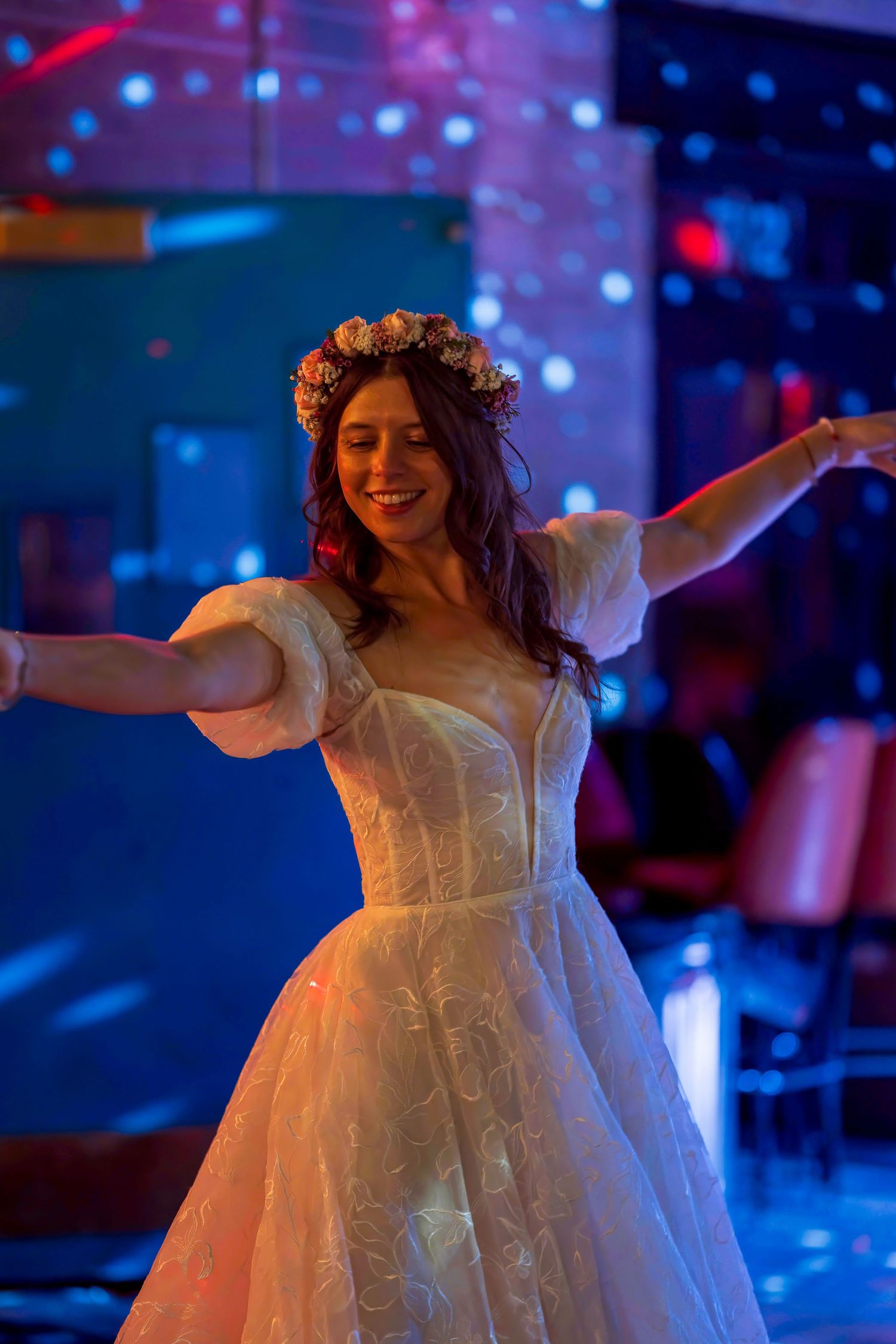 Smiling bride in white lace gown and floral crown dancing joyfully with arms outstretched at a reception.