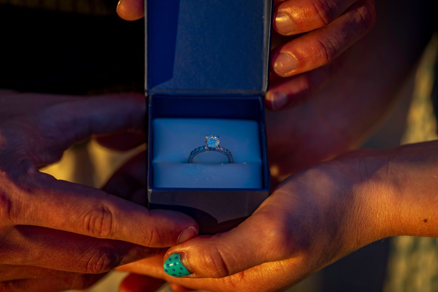 Hands holding an open blue jewelry box containing a sparkling diamond engagement ring during a proposal.