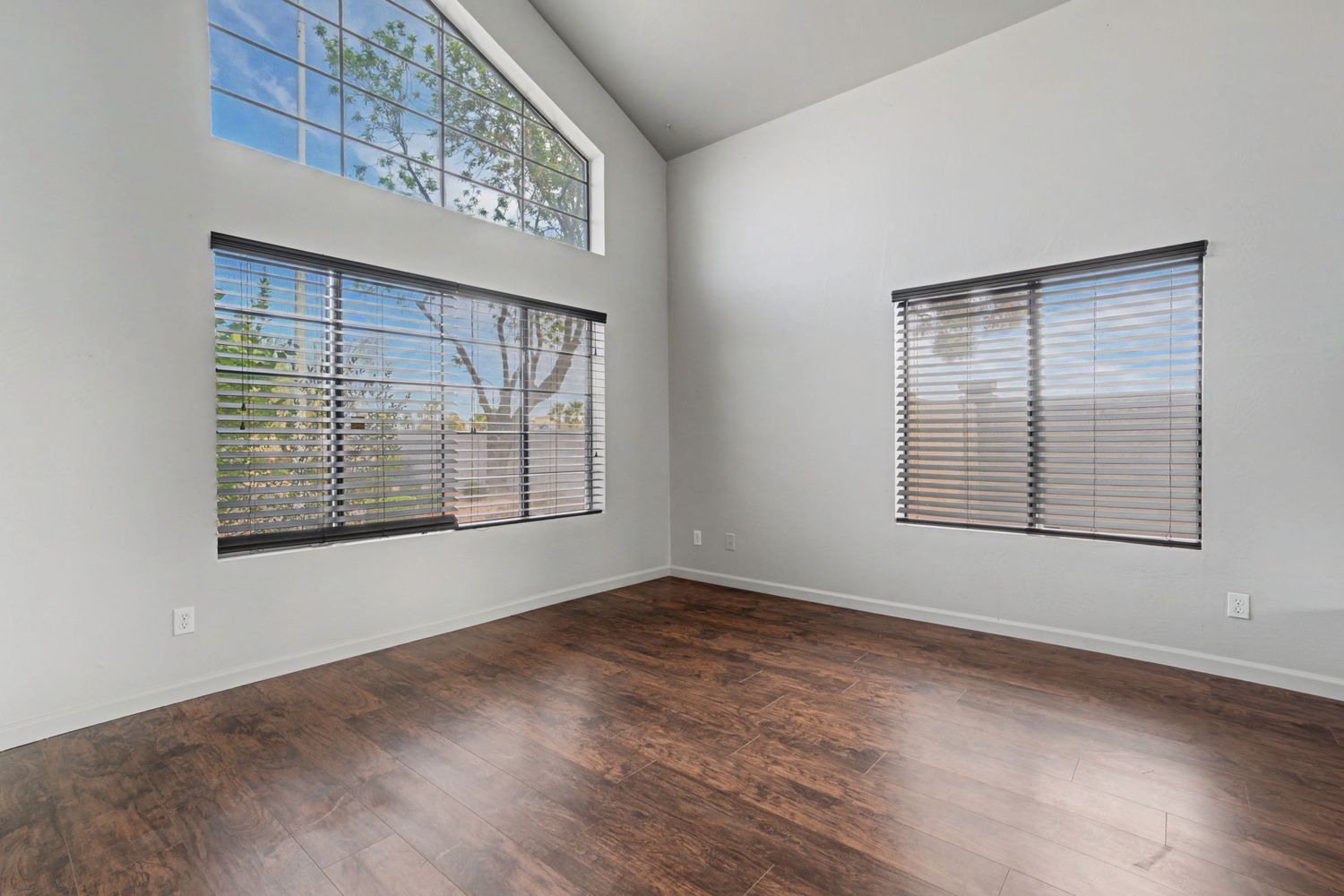 Bright empty room with dark hardwood floors, high ceilings, large windows with blinds, and abundant natural light.