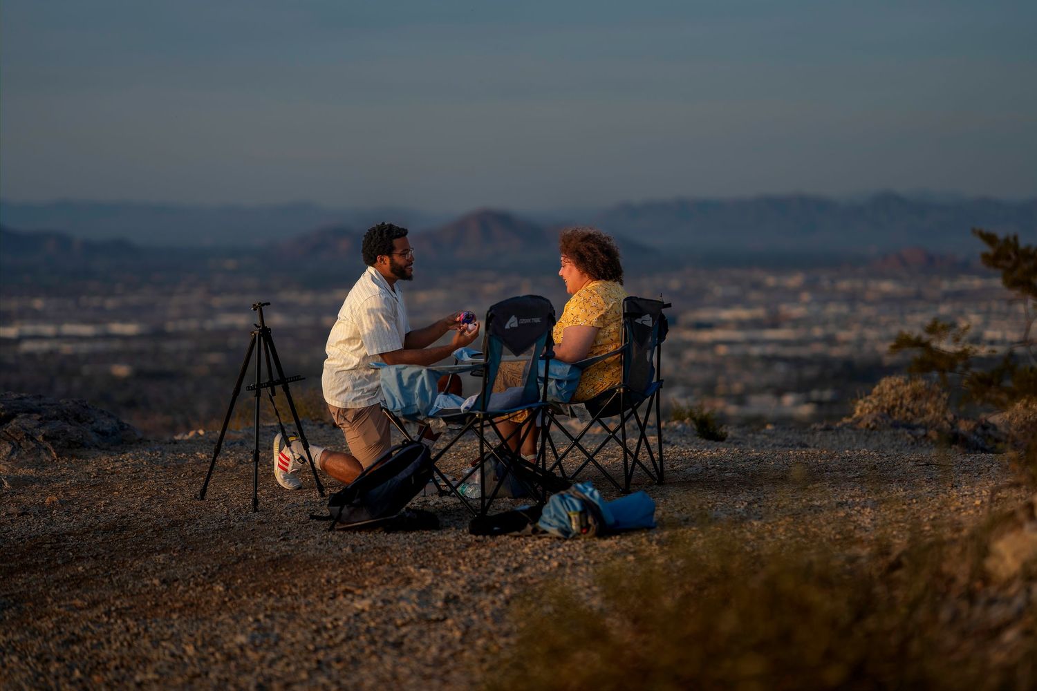 Two people sitting in camping chairs sharing a moment at sunset overlooking a desert cityscape.
