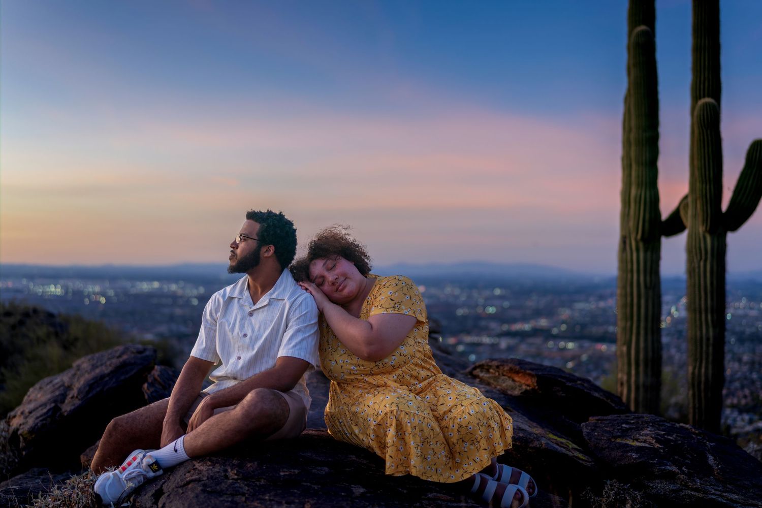 Couple resting on rocks at sunset with saguaro cactus and city view behind them.