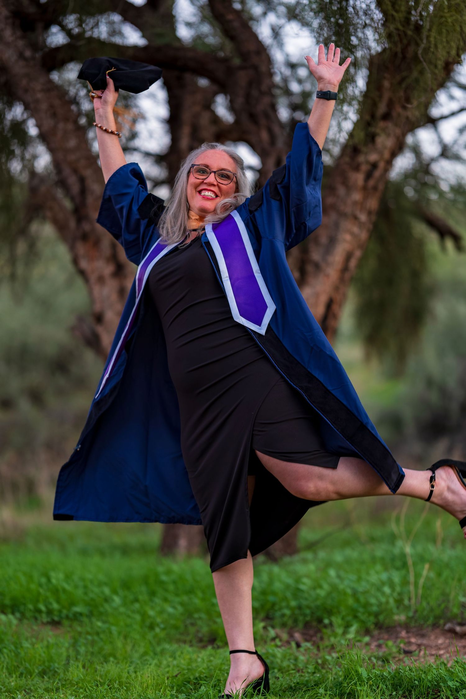 Graduate in blue regalia celebrating with arms raised and cap in hand outdoors.