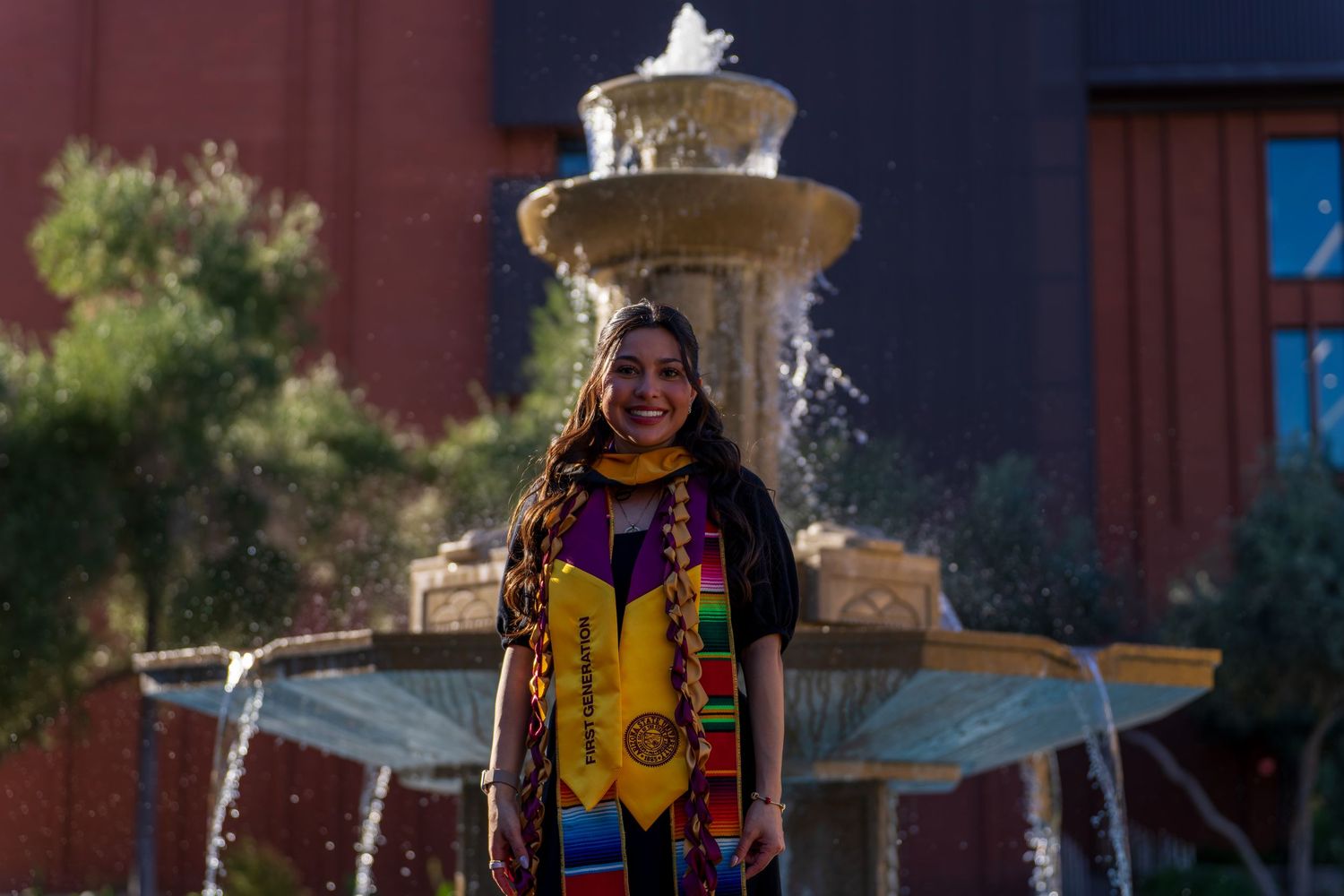 Graduate in traditional regalia poses near fountain on campus.