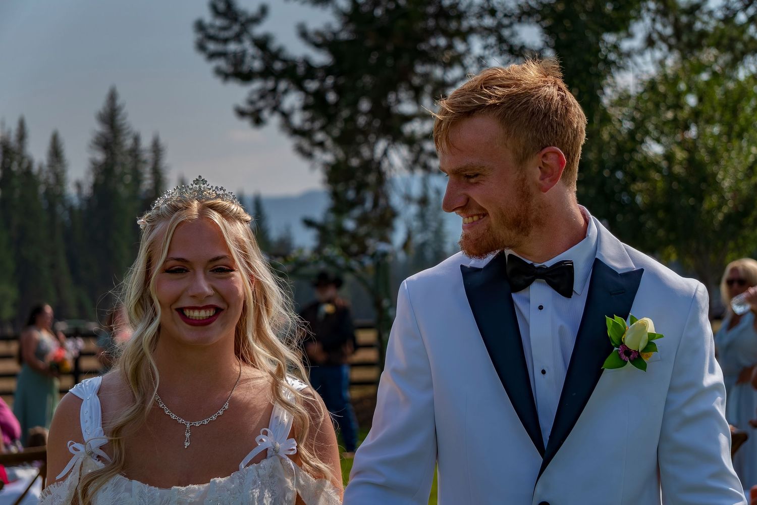 A joyful wedding couple laughs together outdoors with evergreen trees and mountains in the background.