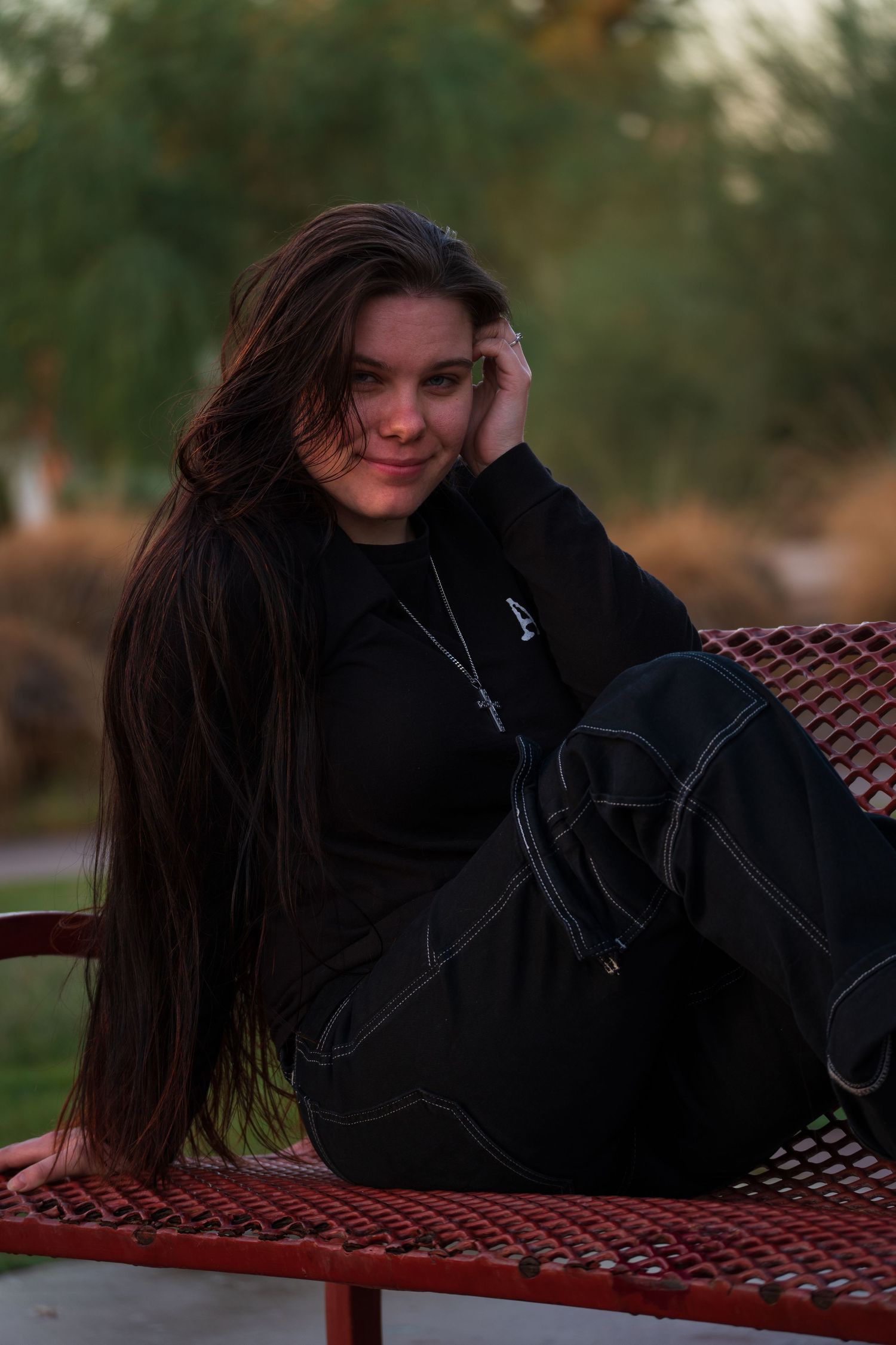 Long dark hair contrasts with black jacket while lounging on red metal bench outdoors.