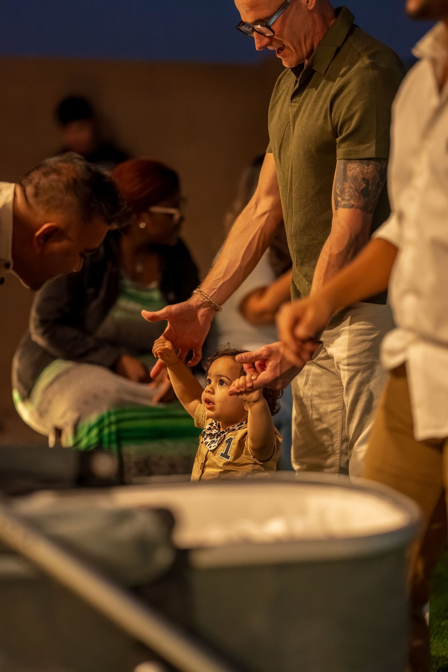 Adult in olive shirt helps small child participate in what appears to be a group activity in warm lighting.