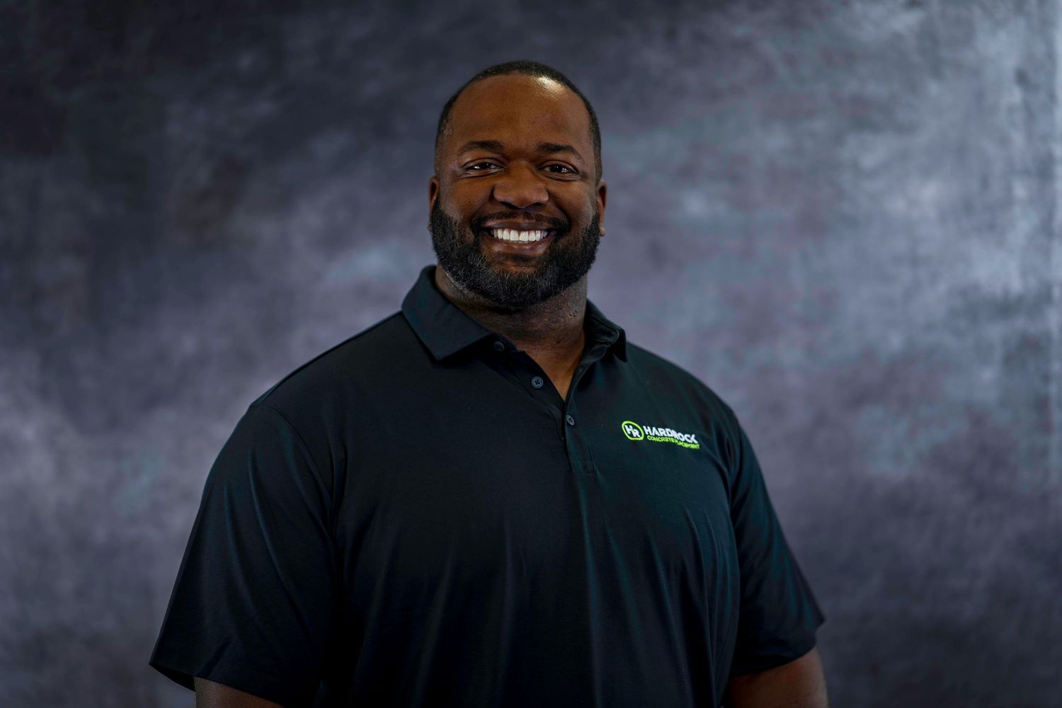 Professional portrait of smiling employee wearing black polo with company logo on gray backdrop.