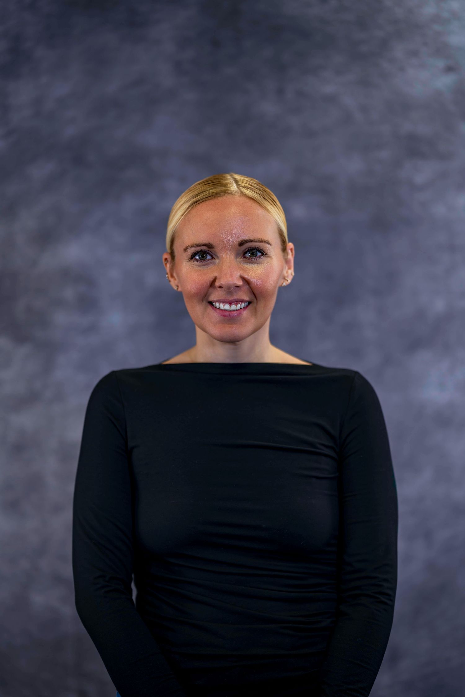 Portrait of someone in a black long-sleeve top smiling against a mottled gray backdrop.