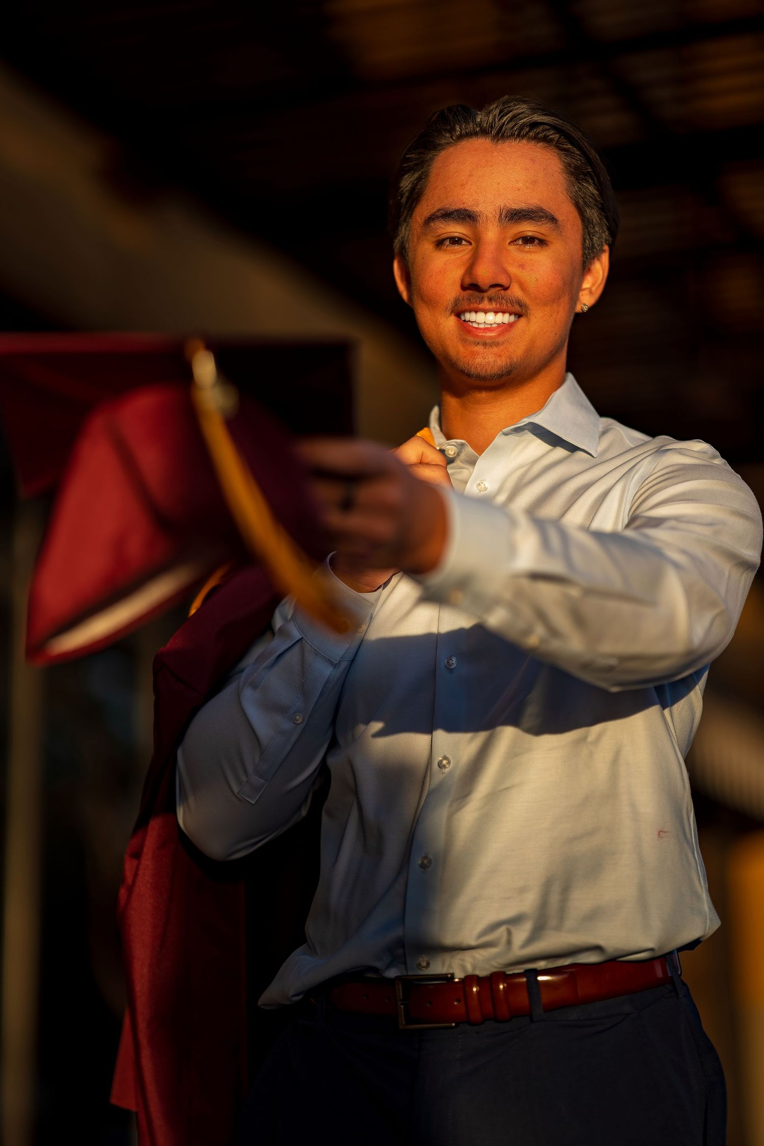 Graduate in maroon gown and light blue shirt celebrating with arms raised.