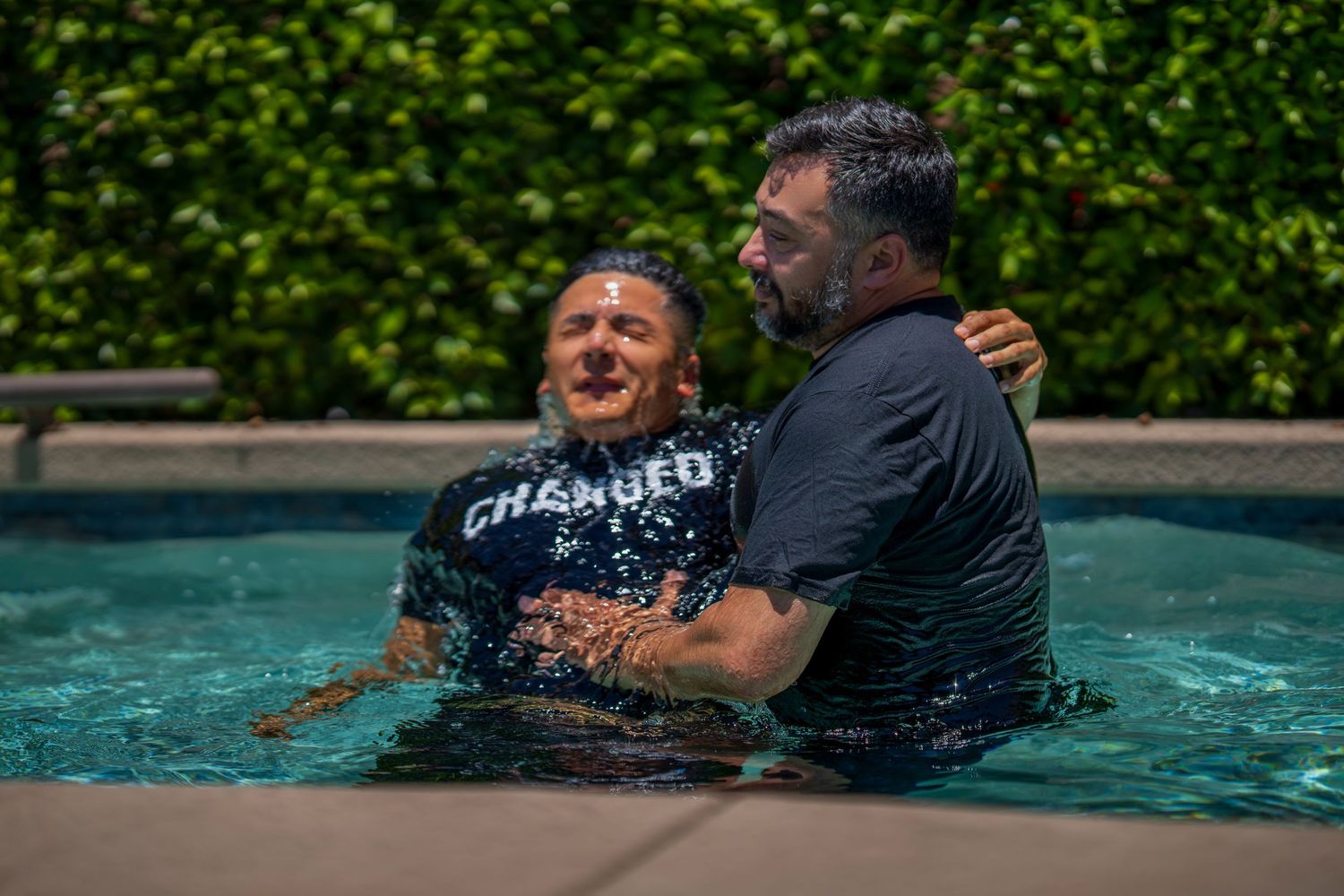 Two men sharing a joyful baptism moment in a pool, one wearing a 'Changed' shirt, surrounded by lush greenery.