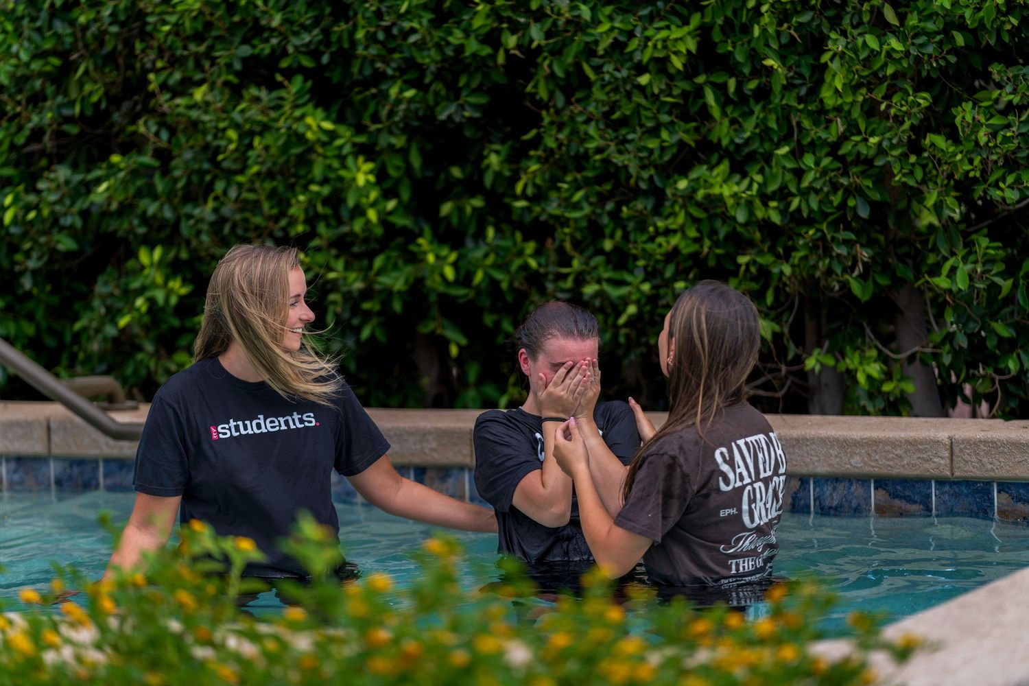 Three people sharing an emotional baptism moment together in an outdoor pool surrounded by greenery.