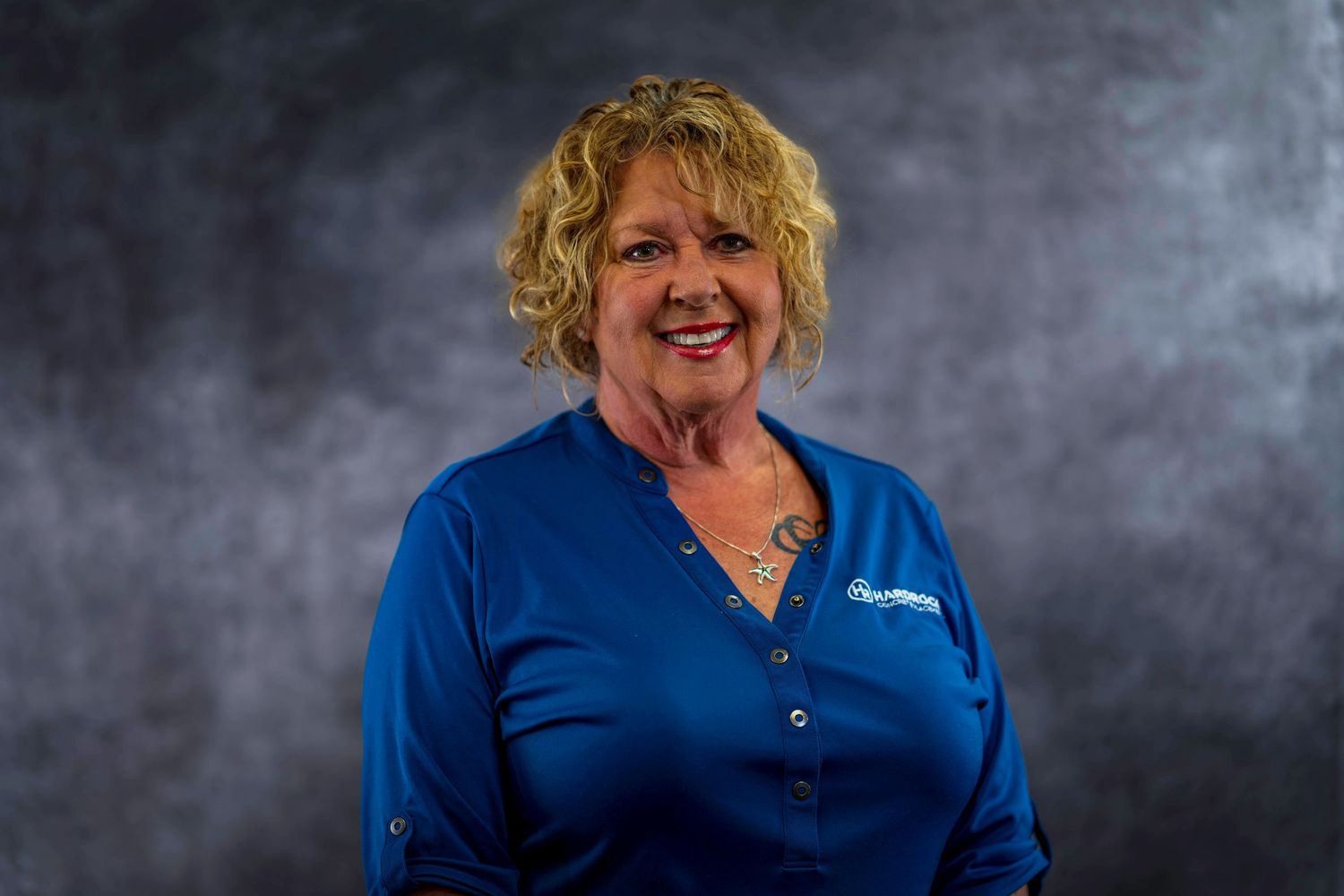 Professional portrait of employee with curly blonde hair wearing blue company shirt against gray backdrop.