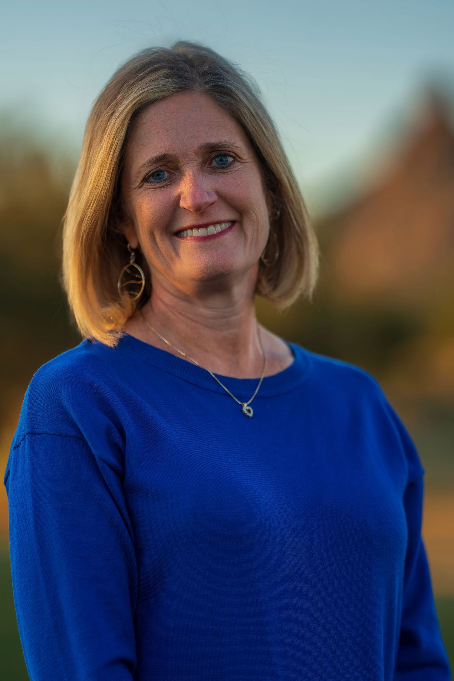 Portrait of person in royal blue sweater with jewelry against sunset backdrop.