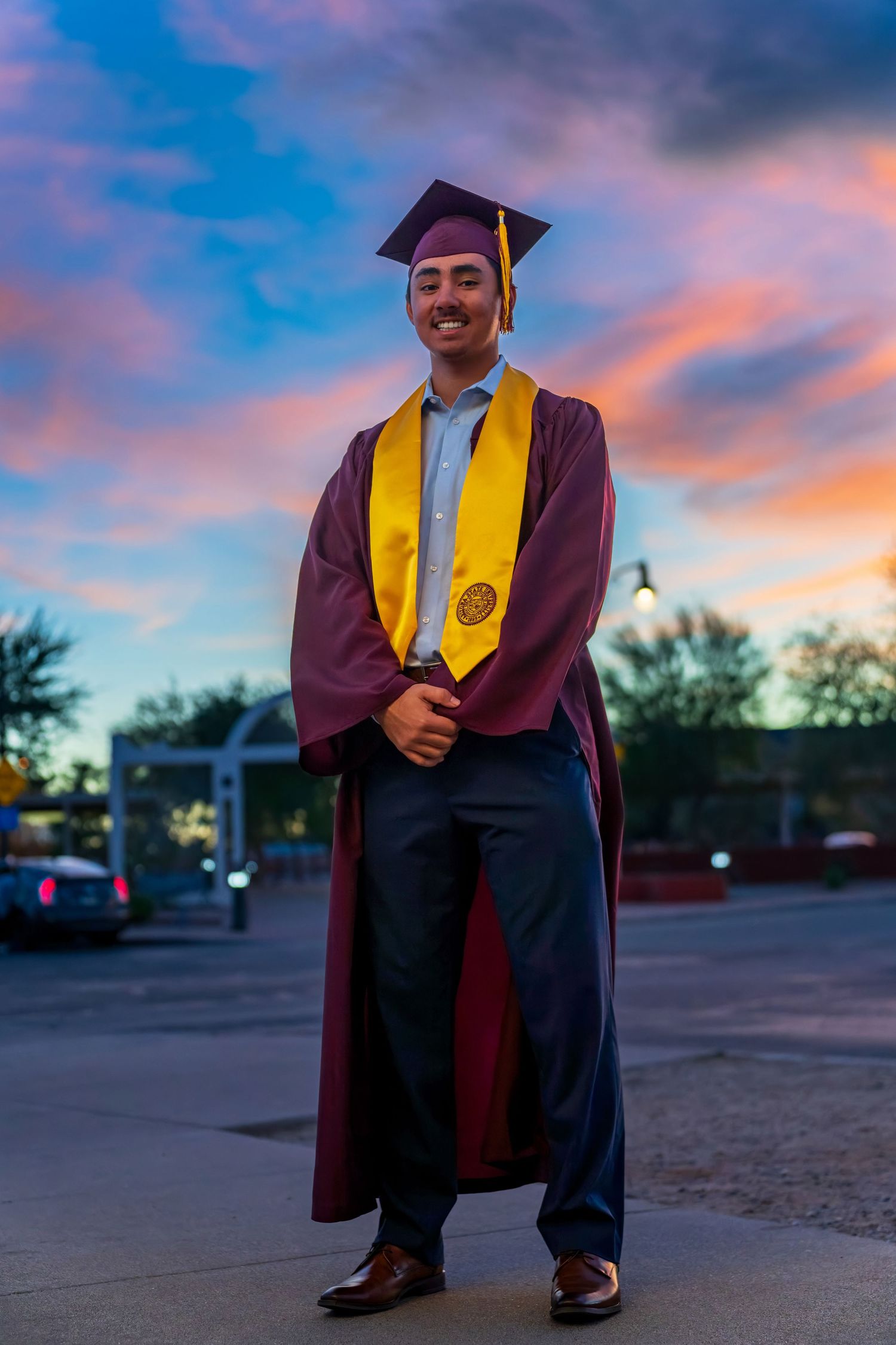 Graduate in maroon and gold regalia poses at sunset in parking lot.