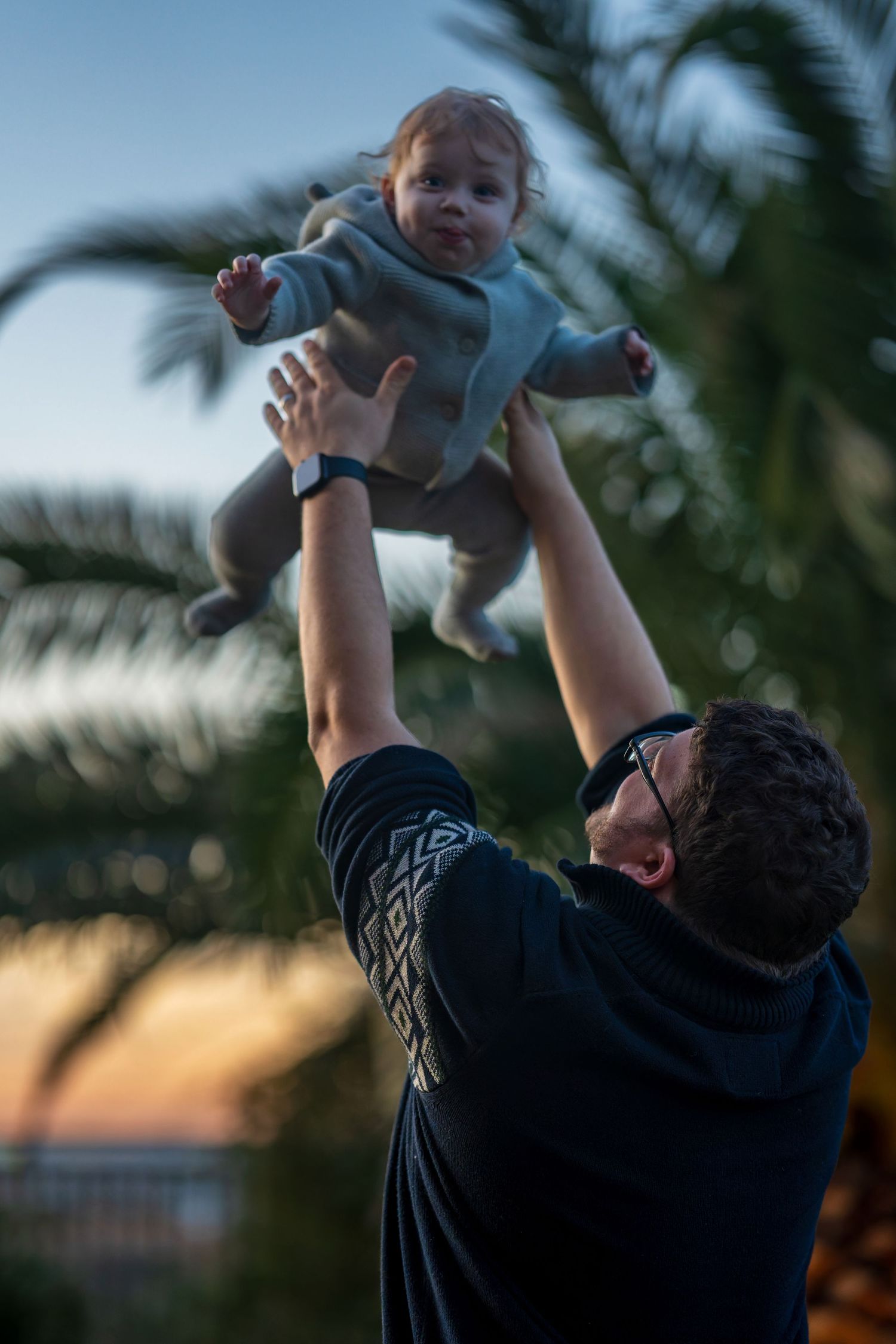Silhouette of person lifting baby high in the air at sunset with palm trees in background.