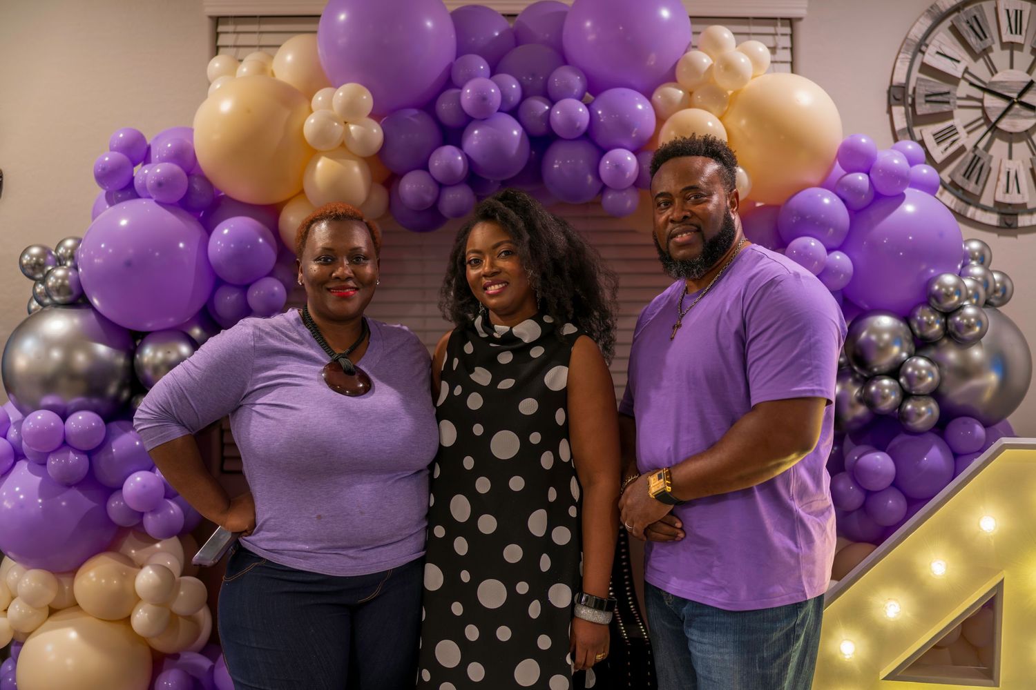 Three people smiling in front of a purple, gold, and silver balloon arch at a festive celebration.