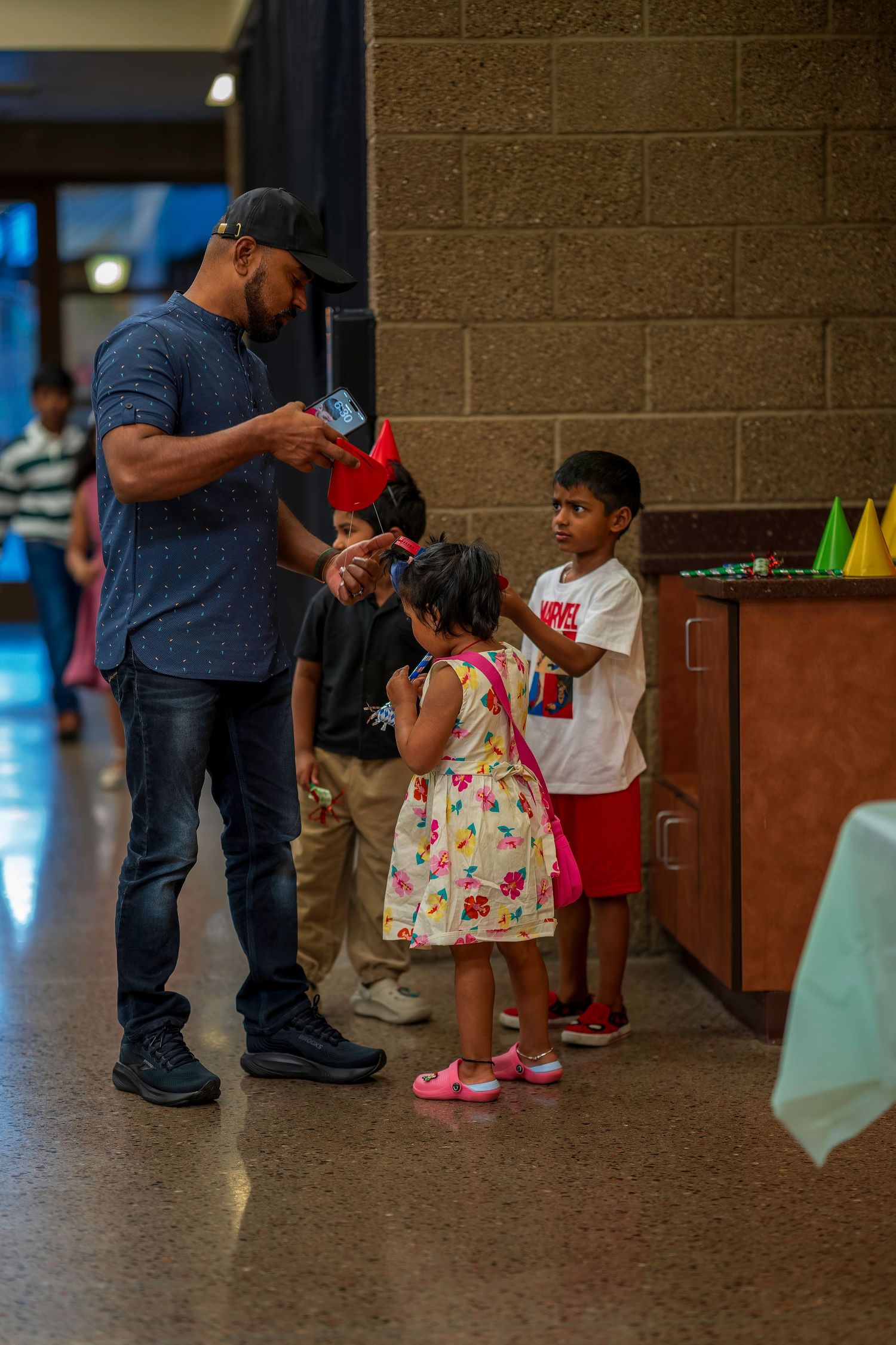 A man in a blue shirt and cap interacts with young children at an indoor birthday party celebration.