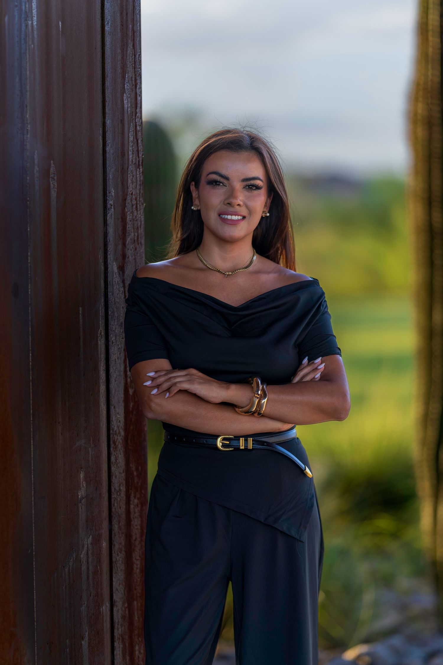 Woman in black off-shoulder top and pants, arms crossed, leaning against wooden post outdoors.
