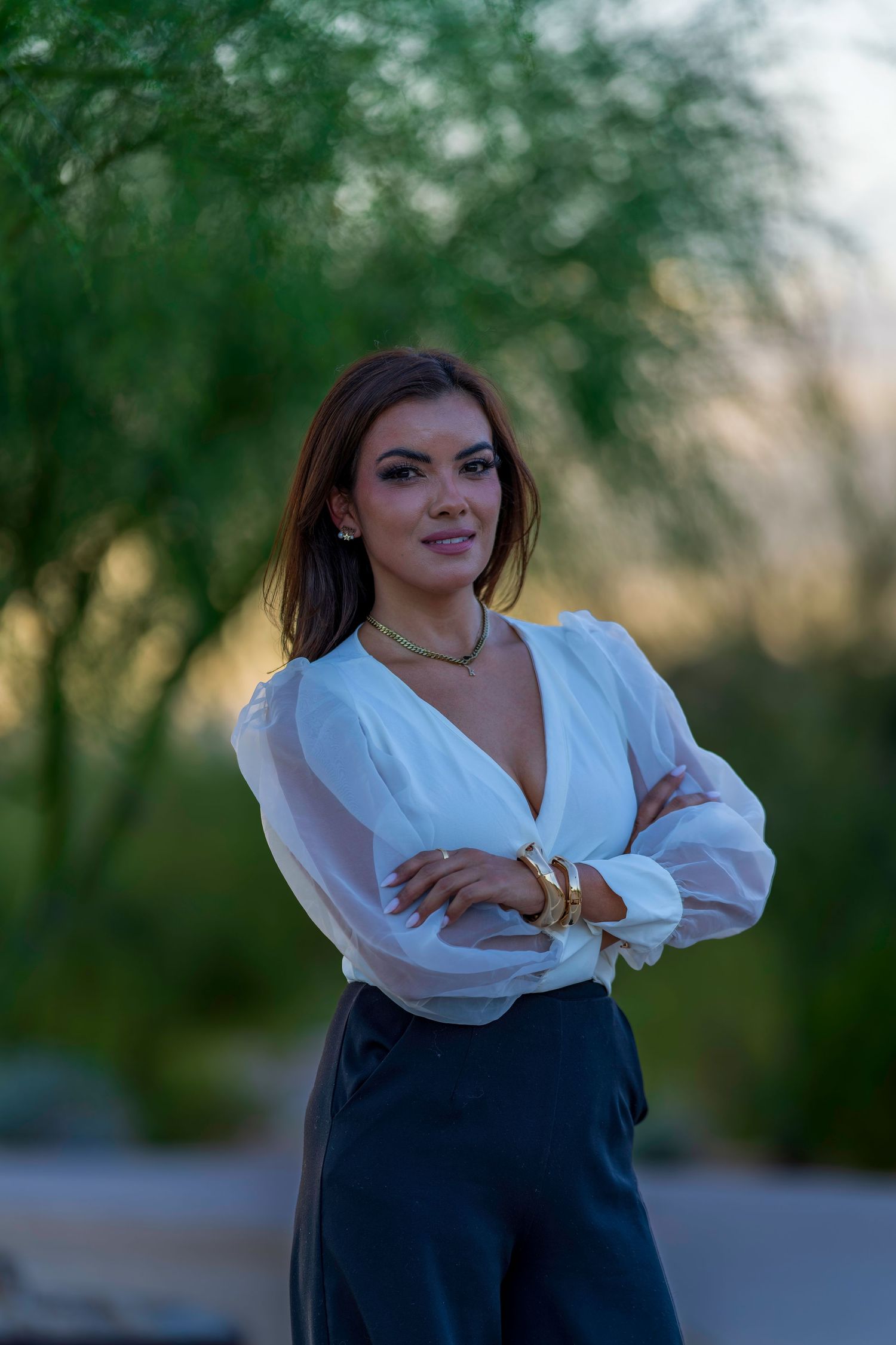 Woman in white blouse with arms crossed smiling outdoors at sunset with green trees in background.