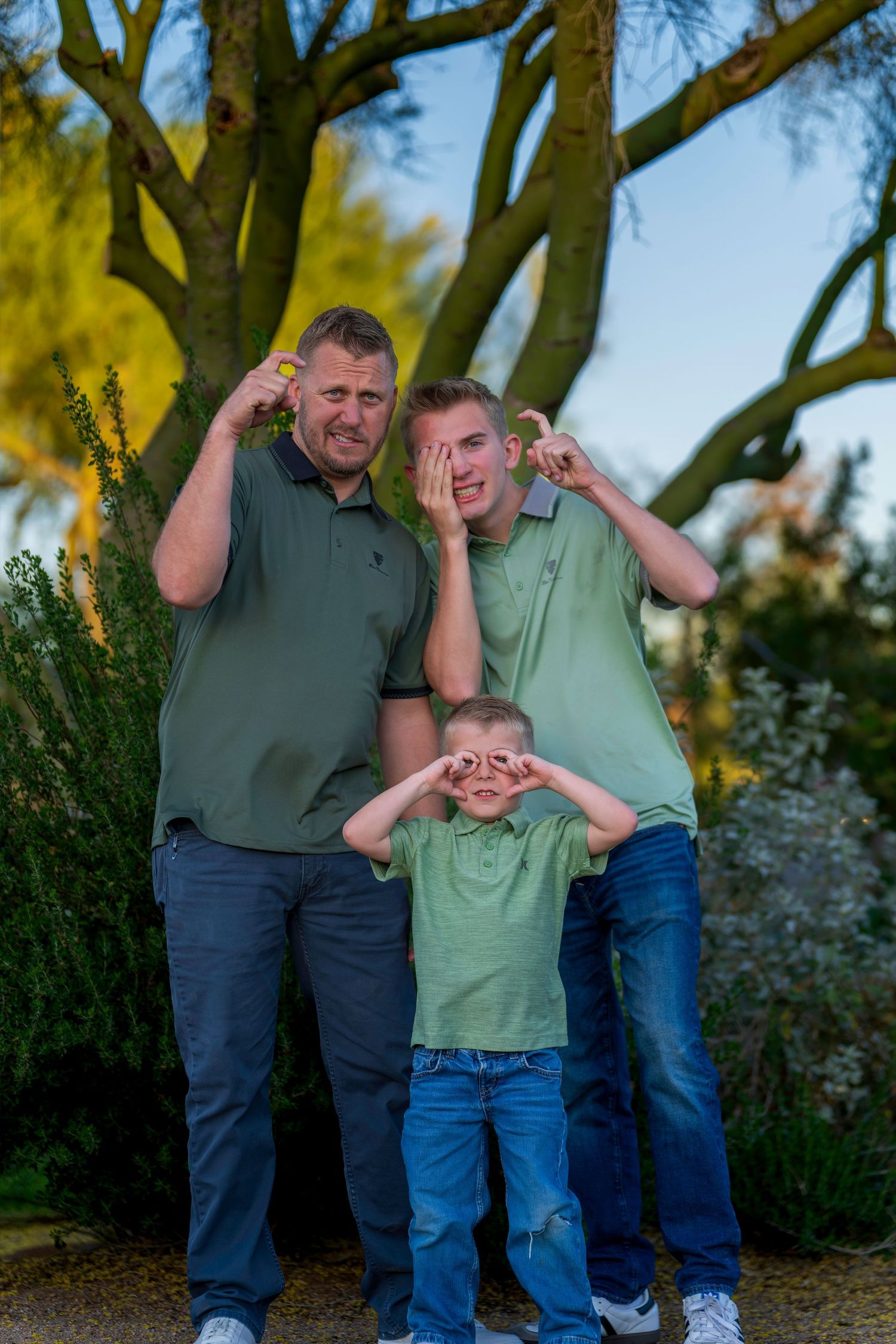 Three males in matching green polo shirts make silly faces outdoors with trees in background.