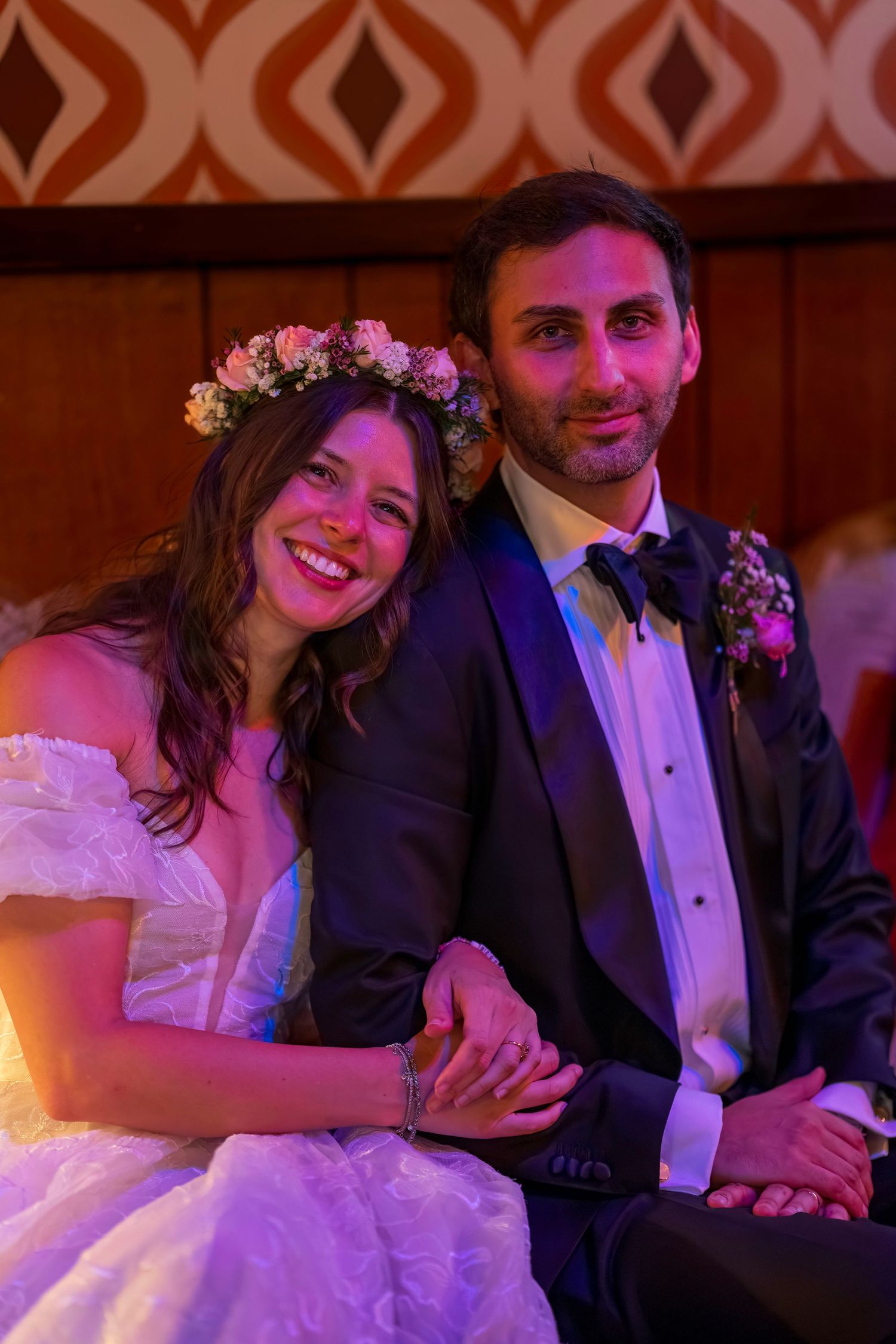Smiling bride in floral crown and groom in navy suit holding hands at wedding reception.