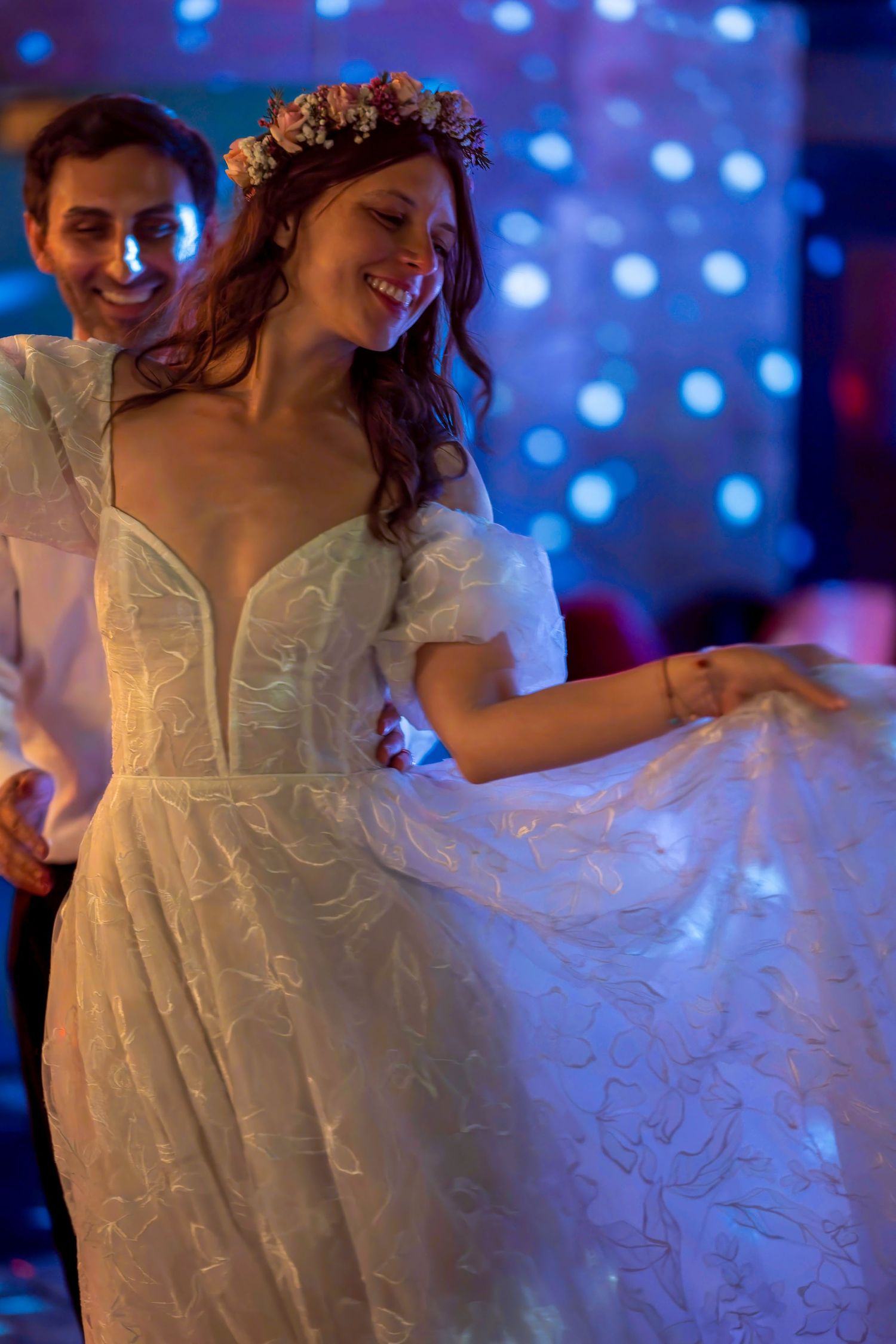 Bride in white lace gown with floral crown dancing joyfully with groom at wedding reception.