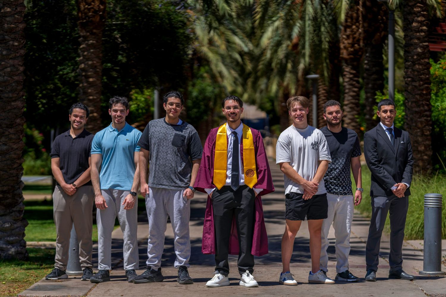 Graduate in maroon cap and gown poses with friends outdoors on tree-lined campus path.